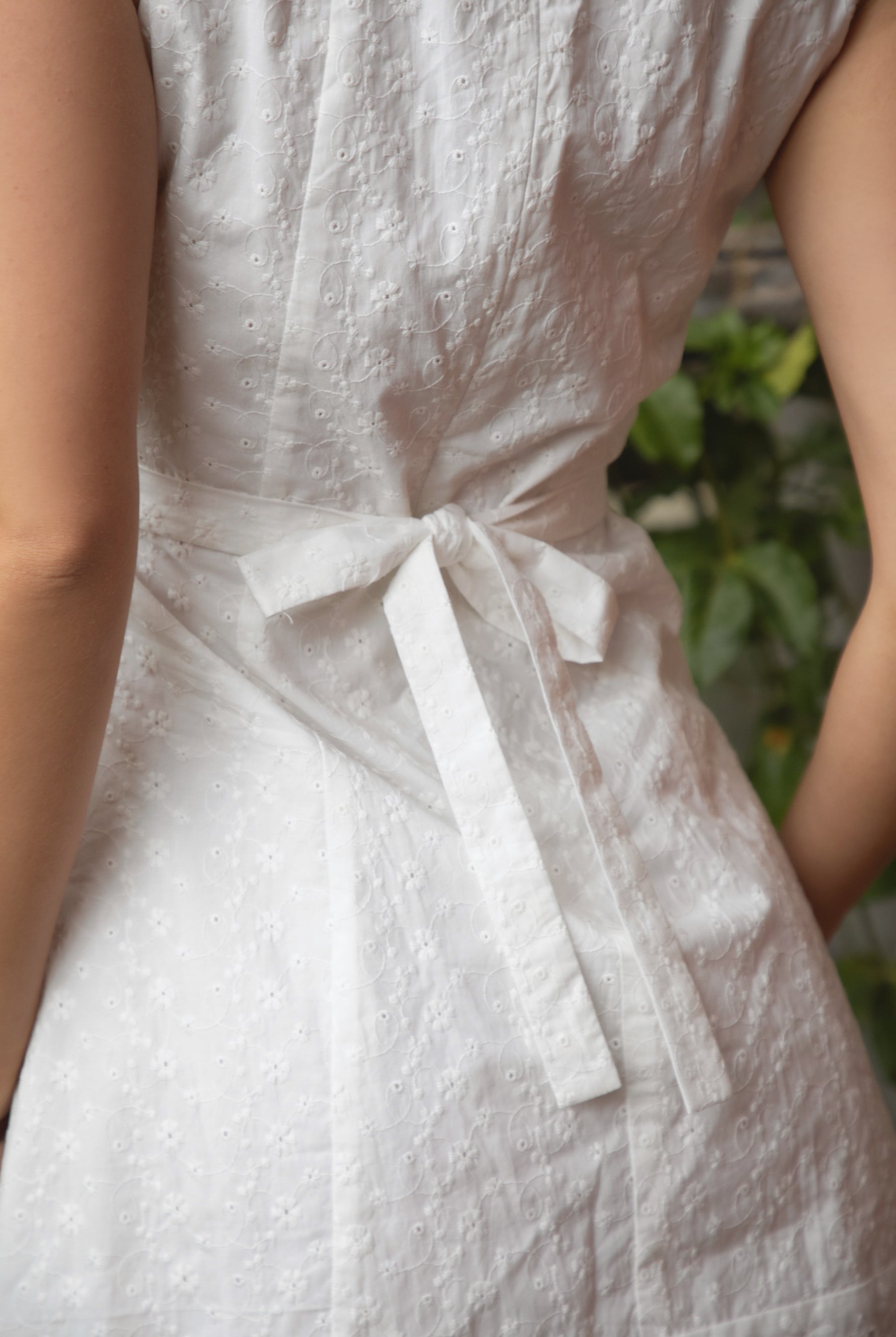 Woman wearing TIC TOC DA31929-AMAYRA AMAYRA eyelet mini dress made from allover eyelet embroidered cotton fabric, featuring a sleeveless square neckline, faux button front detail, fitted bodice with clean seams, adjustable back waist tie, hidden back zipper, and scalloped eyelet lace hemline, styled for a brunch-ready garden party look.