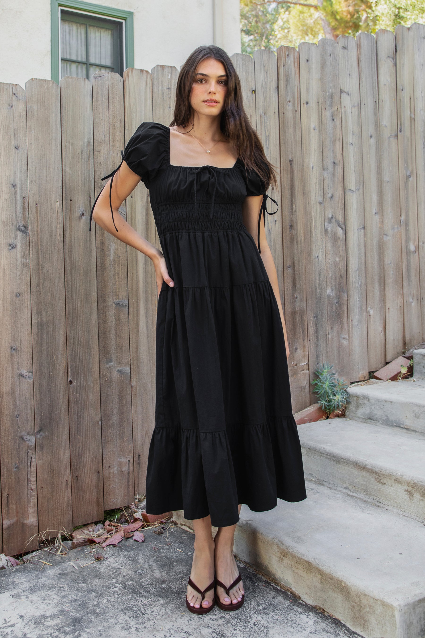 Woman wearing WYNORA smocked puff sleeve tiered midi dress featuring smooth stretch poplin fabric, soft jersey lining, square neckline with adjustable front tie detail, smocked bodice, short puff sleeves with ribbon tie accents, and a flowing tiered midi skirt with ruffle hem finish.