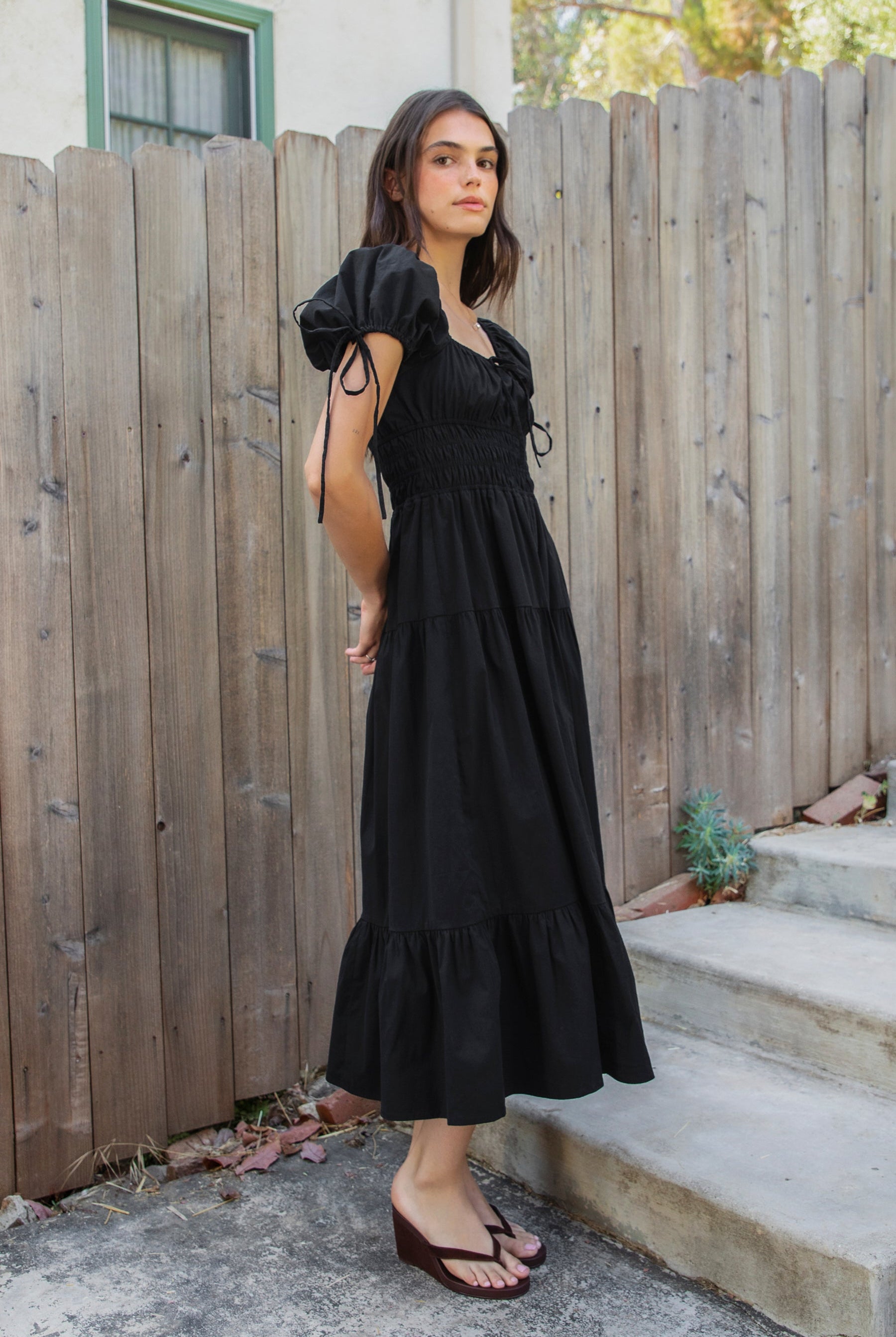 Woman wearing WYNORA smocked puff sleeve tiered midi dress featuring smooth stretch poplin fabric, soft jersey lining, square neckline with adjustable front tie detail, smocked bodice, short puff sleeves with ribbon tie accents, and a flowing tiered midi skirt with ruffle hem finish.