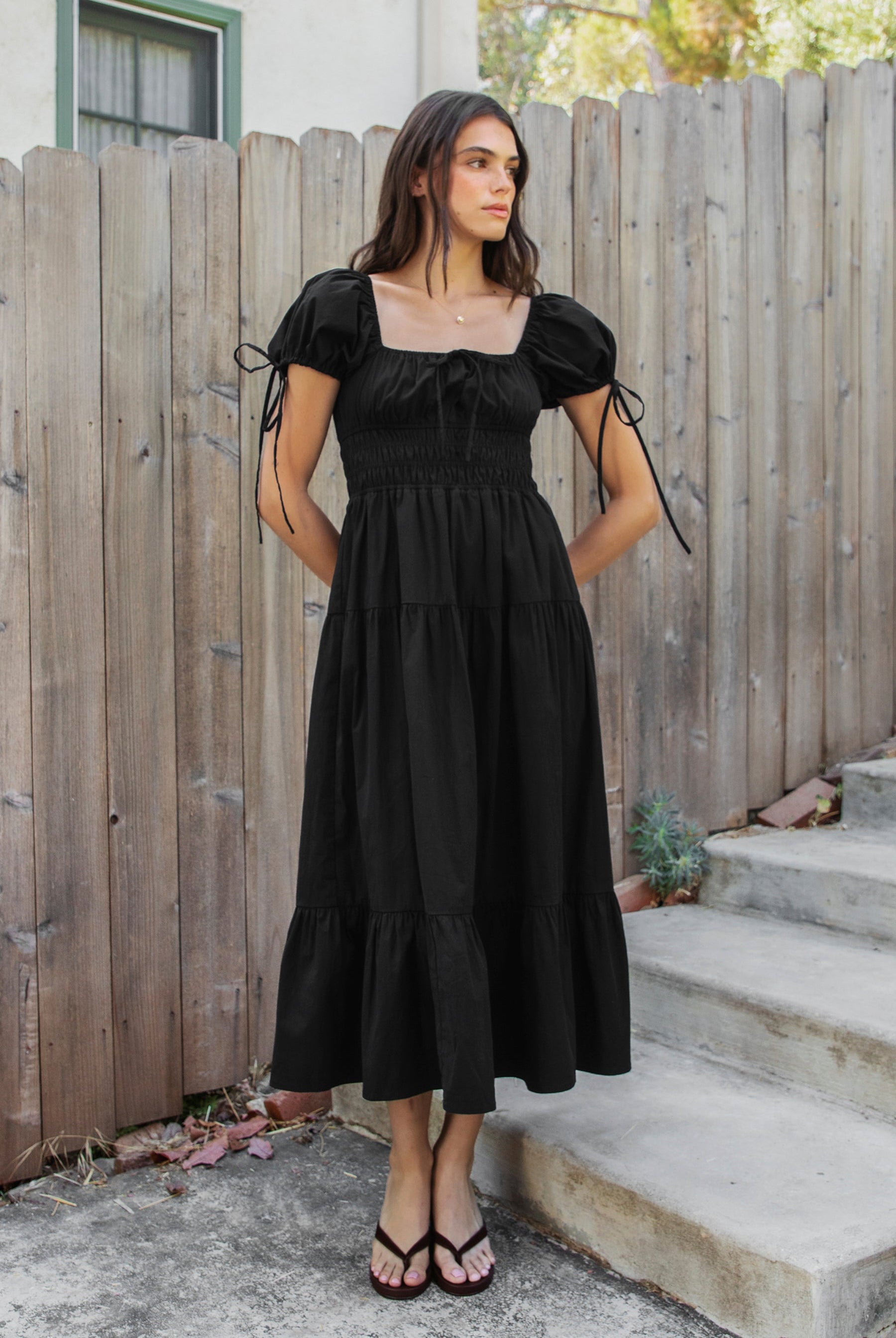 Woman wearing WYNORA smocked puff sleeve tiered midi dress featuring smooth stretch poplin fabric, soft jersey lining, square neckline with adjustable front tie detail, smocked bodice, short puff sleeves with ribbon tie accents, and a flowing tiered midi skirt with ruffle hem finish.