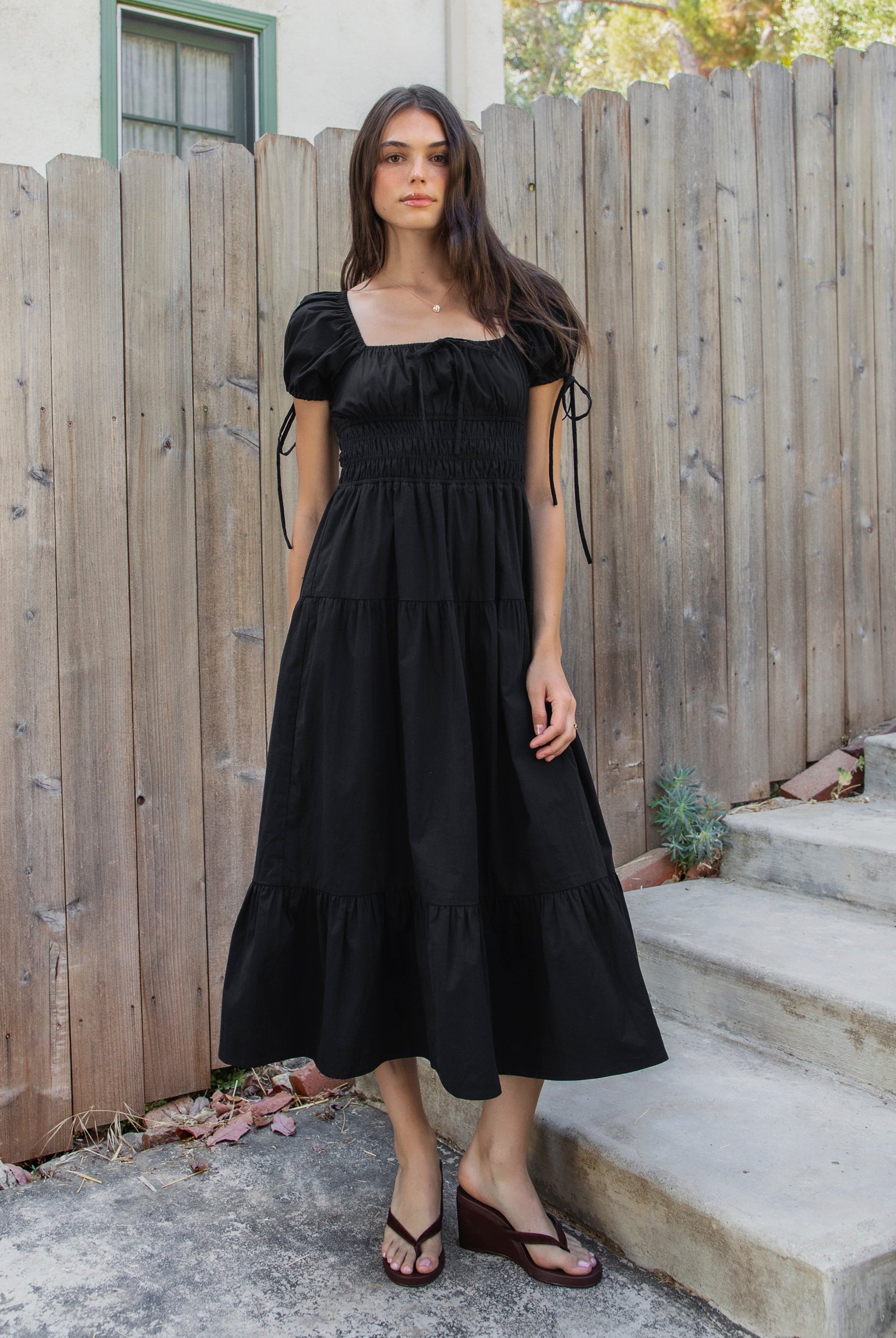Woman wearing WYNORA smocked puff sleeve tiered midi dress featuring smooth stretch poplin fabric, soft jersey lining, square neckline with adjustable front tie detail, smocked bodice, short puff sleeves with ribbon tie accents, and a flowing tiered midi skirt with ruffle hem finish.