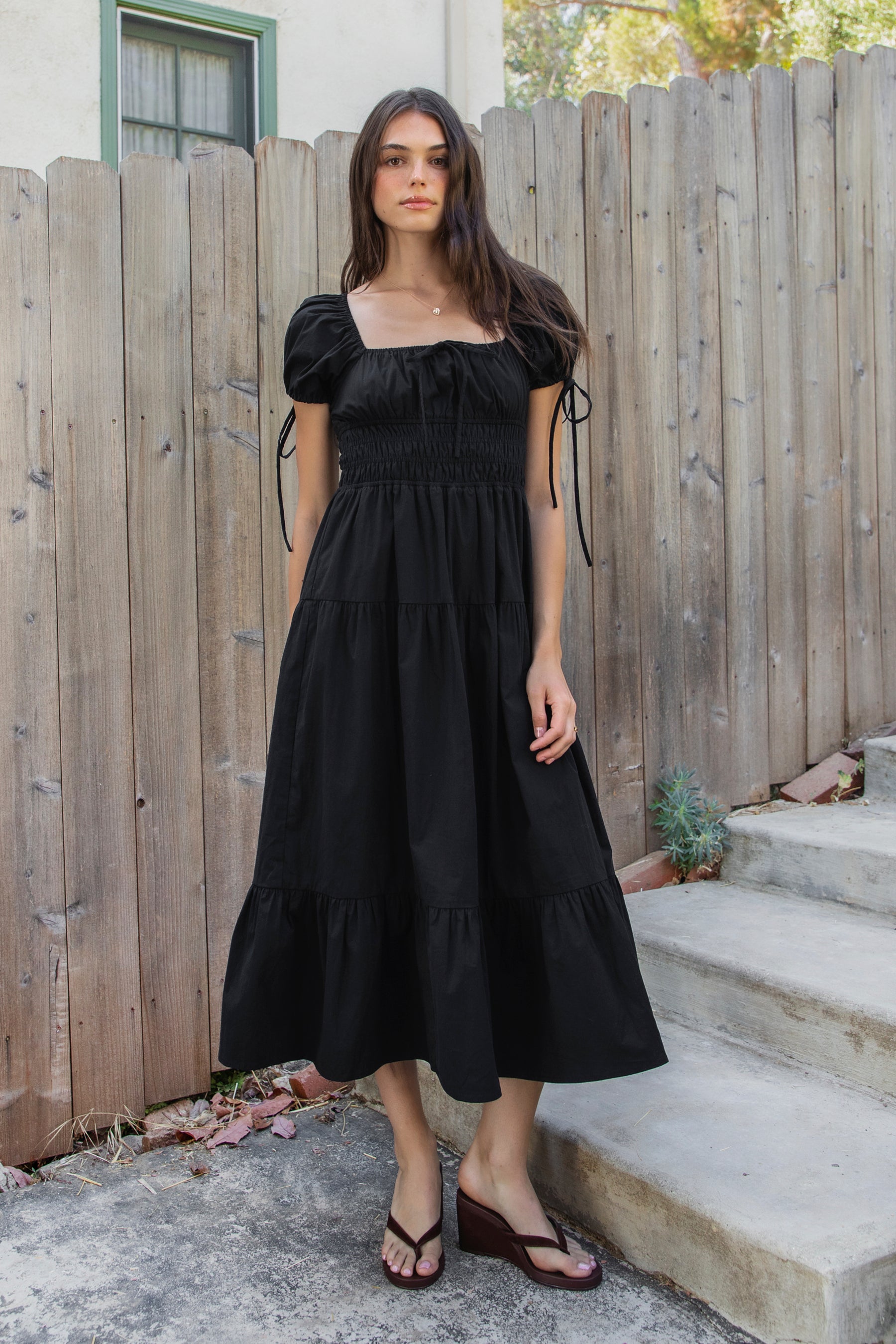 Woman wearing WYNORA smocked puff sleeve tiered midi dress featuring smooth stretch poplin fabric, soft jersey lining, square neckline with adjustable front tie detail, smocked bodice, short puff sleeves with ribbon tie accents, and a flowing tiered midi skirt with ruffle hem finish.