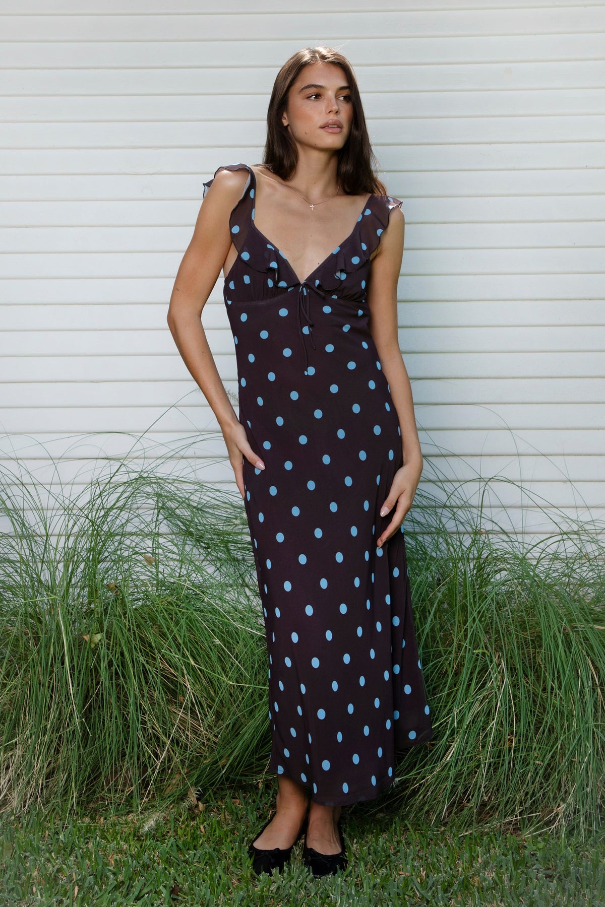 Woman wearing NOIRA ruffle chiffon polka dot maxi dress featuring flowy chiffon fabric, contrast polka dot print, deep V neckline with feminine ruffle trim, ruffled shoulder straps, adjustable front tie detail at the bust, fitted empire waist silhouette, low open back, adjustable shoulder straps, and soft stretch satin lining.