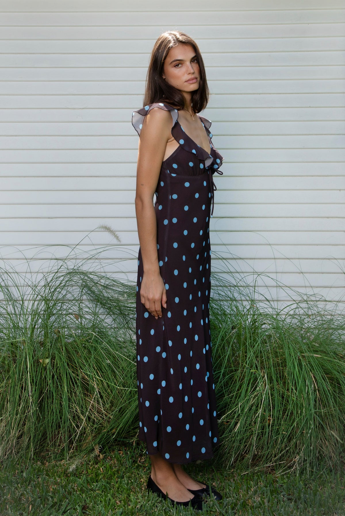 Woman wearing NOIRA ruffle chiffon polka dot maxi dress featuring flowy chiffon fabric, contrast polka dot print, deep V neckline with feminine ruffle trim, ruffled shoulder straps, adjustable front tie detail at the bust, fitted empire waist silhouette, low open back, adjustable shoulder straps, and soft stretch satin lining.