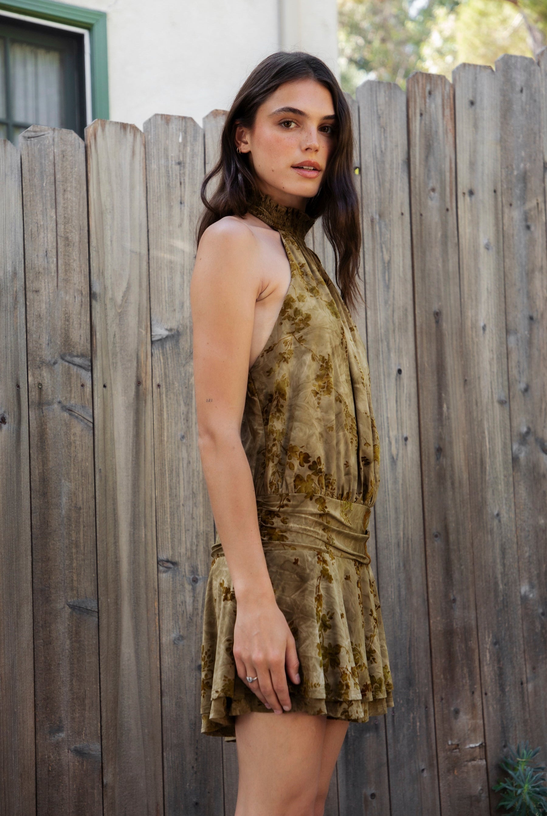 Woman wearing OLIVINE flocked sleeveless floral halter ruched mini dress featuring soft flocked jersey fabric, stretch jersey lining, smocked high halter neckline, open back design, ruched drop waist, layered flounce mini skirt, and pull on construction for special occasion styling.