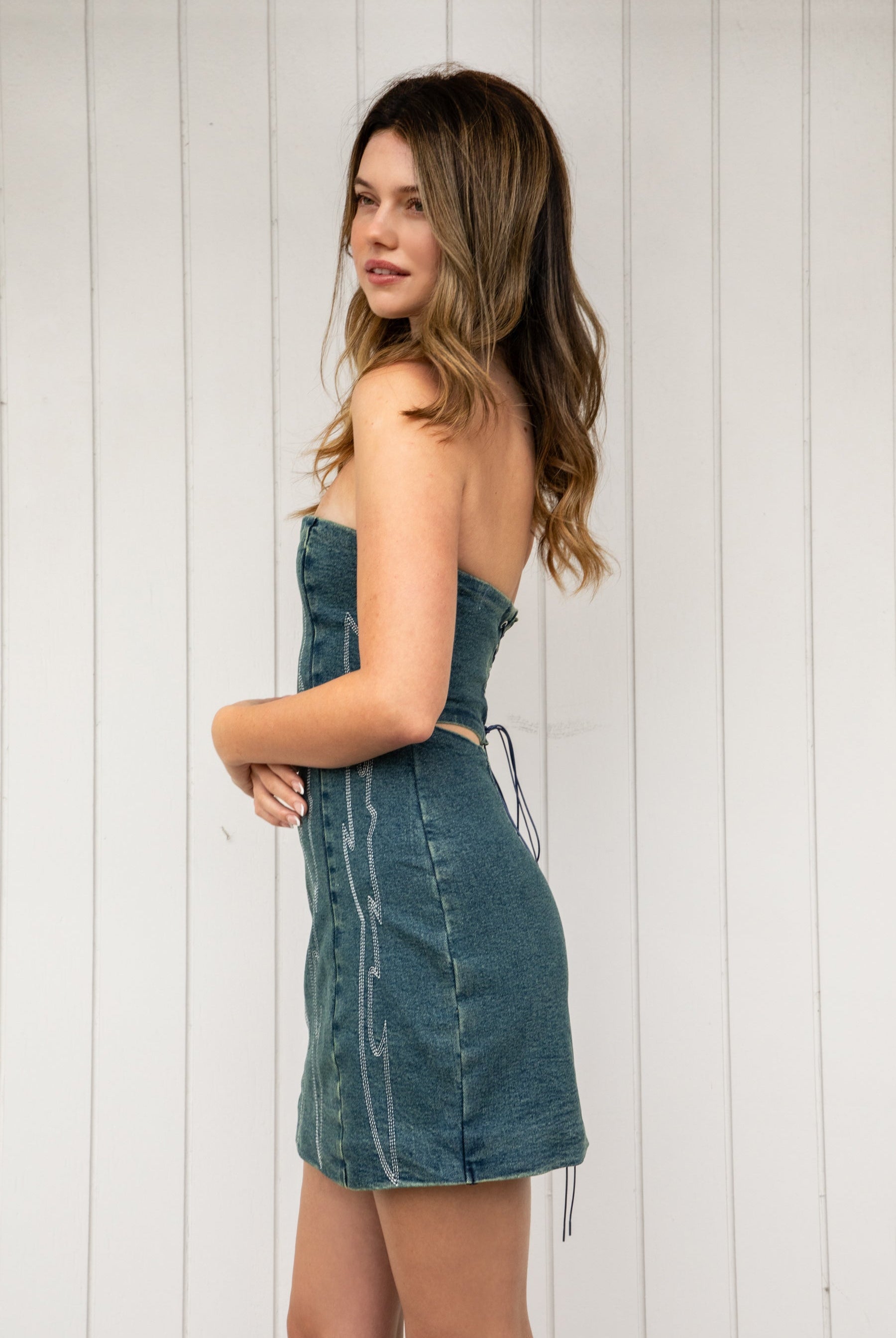 Woman wearing ARIZONA denim western stitch mini dress featuring vintage washed denim, bold western inspired contrast stitching, strapless corset style bodice, lace up back with adjustable closure, subtle back cutout, fitted waist, and a soft stretch mini length silhouette.