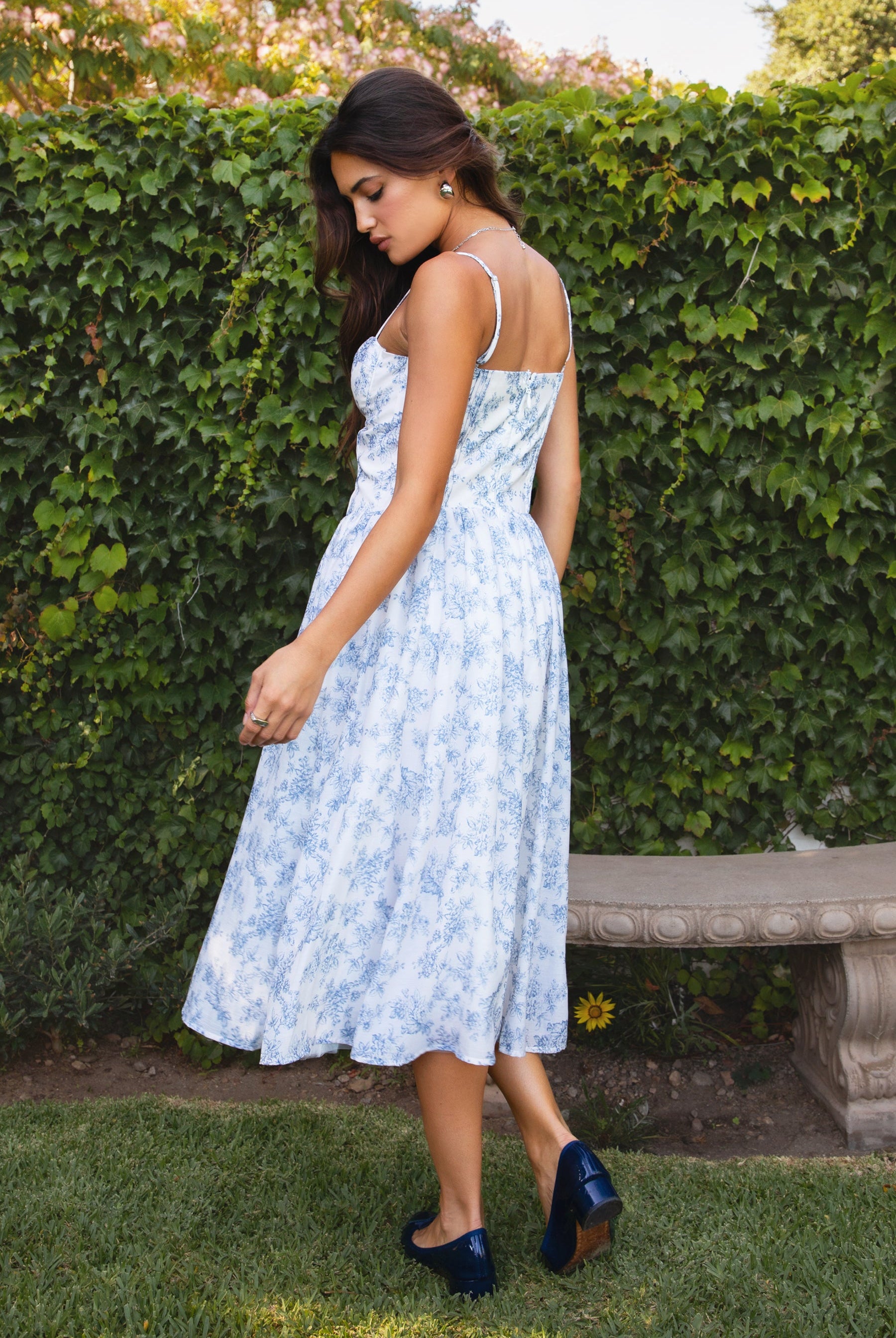 Woman in a light blue floral dress standing in front of a green hedge.