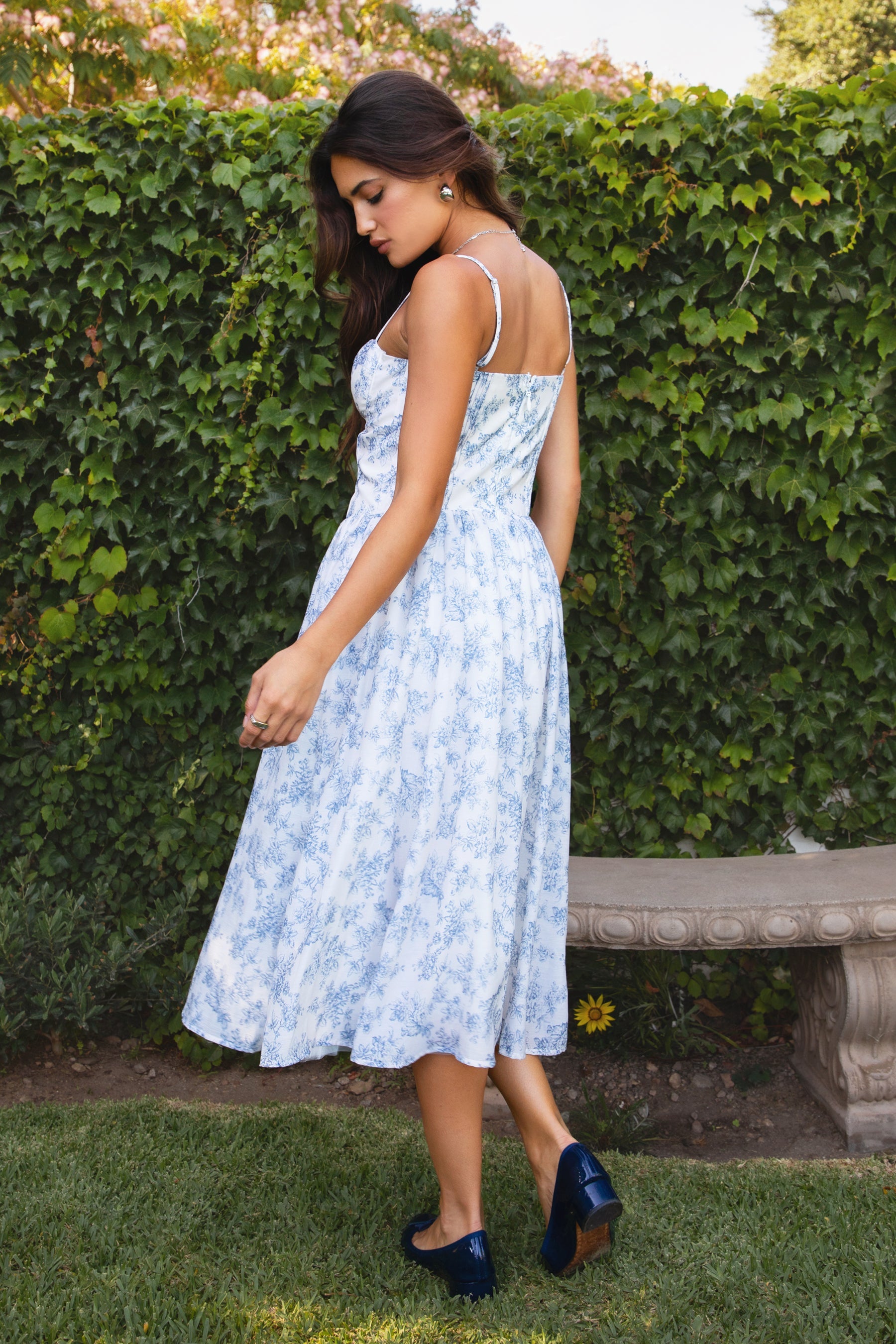Woman in a light blue floral dress standing in front of a green hedge.