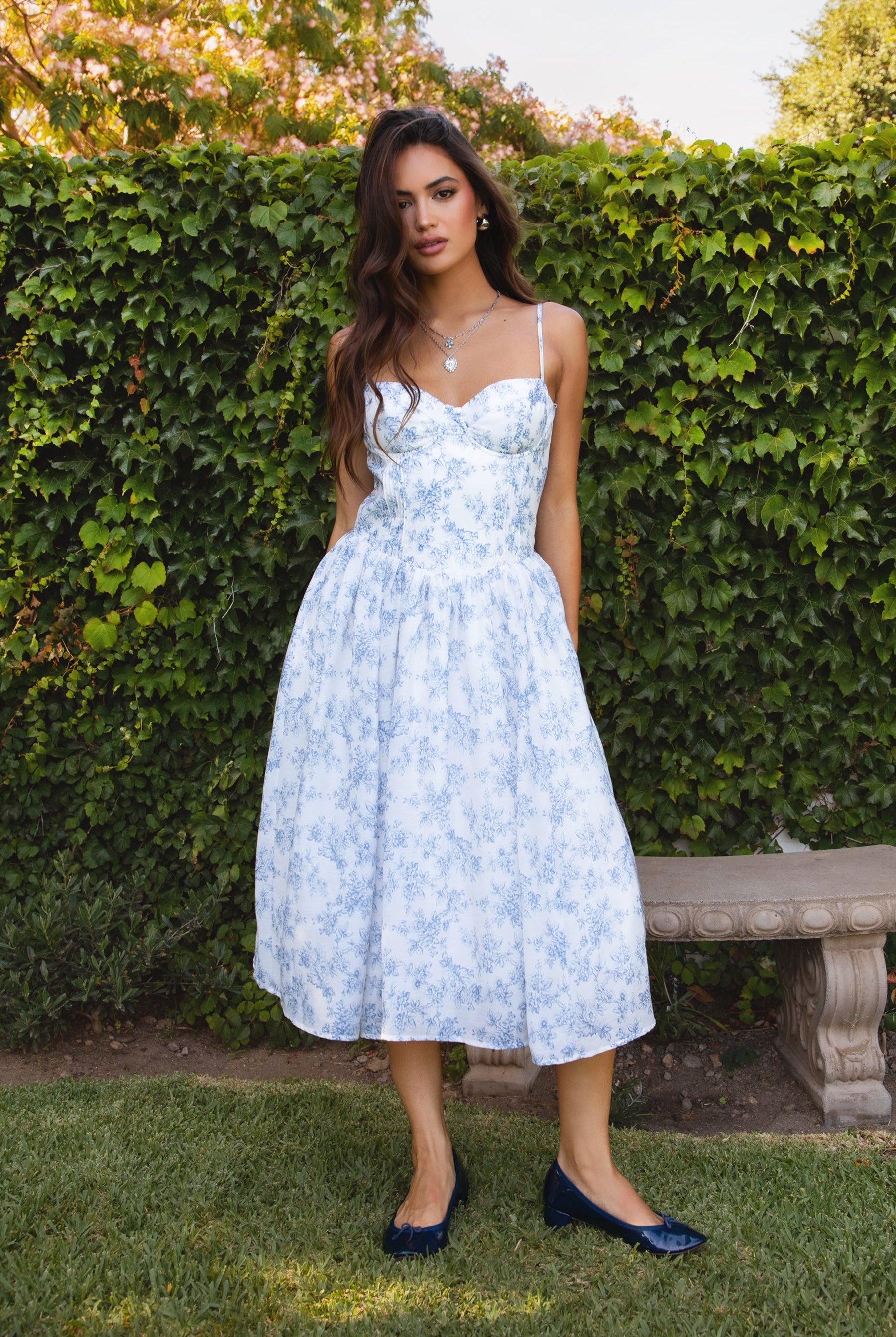 Woman in a light blue floral dress standing in front of a green hedge.
