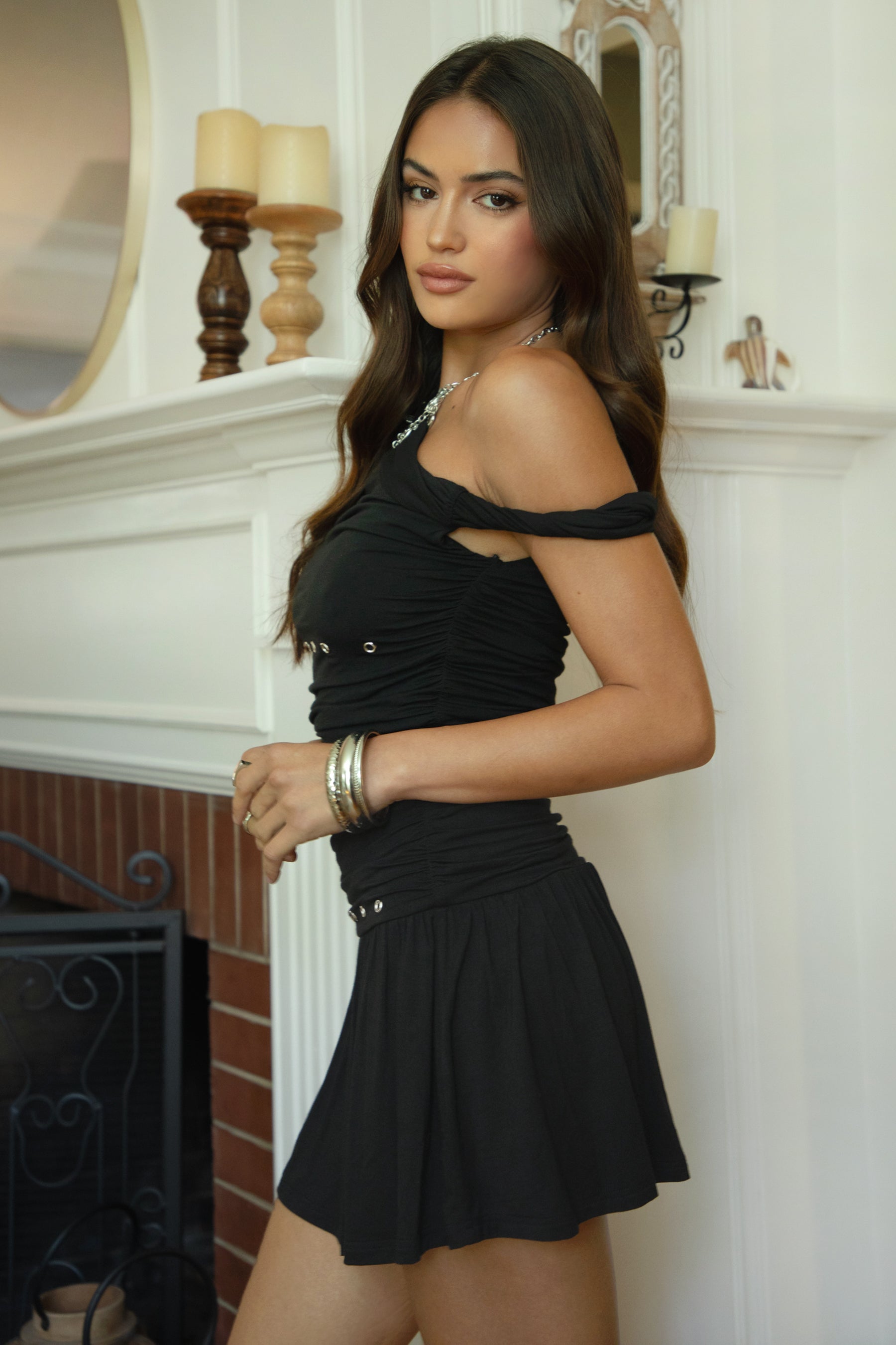 Women wearing TIC TOC AZALEA One Shoulder Mini Dress in black with twisted strap, ruching, studded detail, and flirty hemline in doors near a fire place