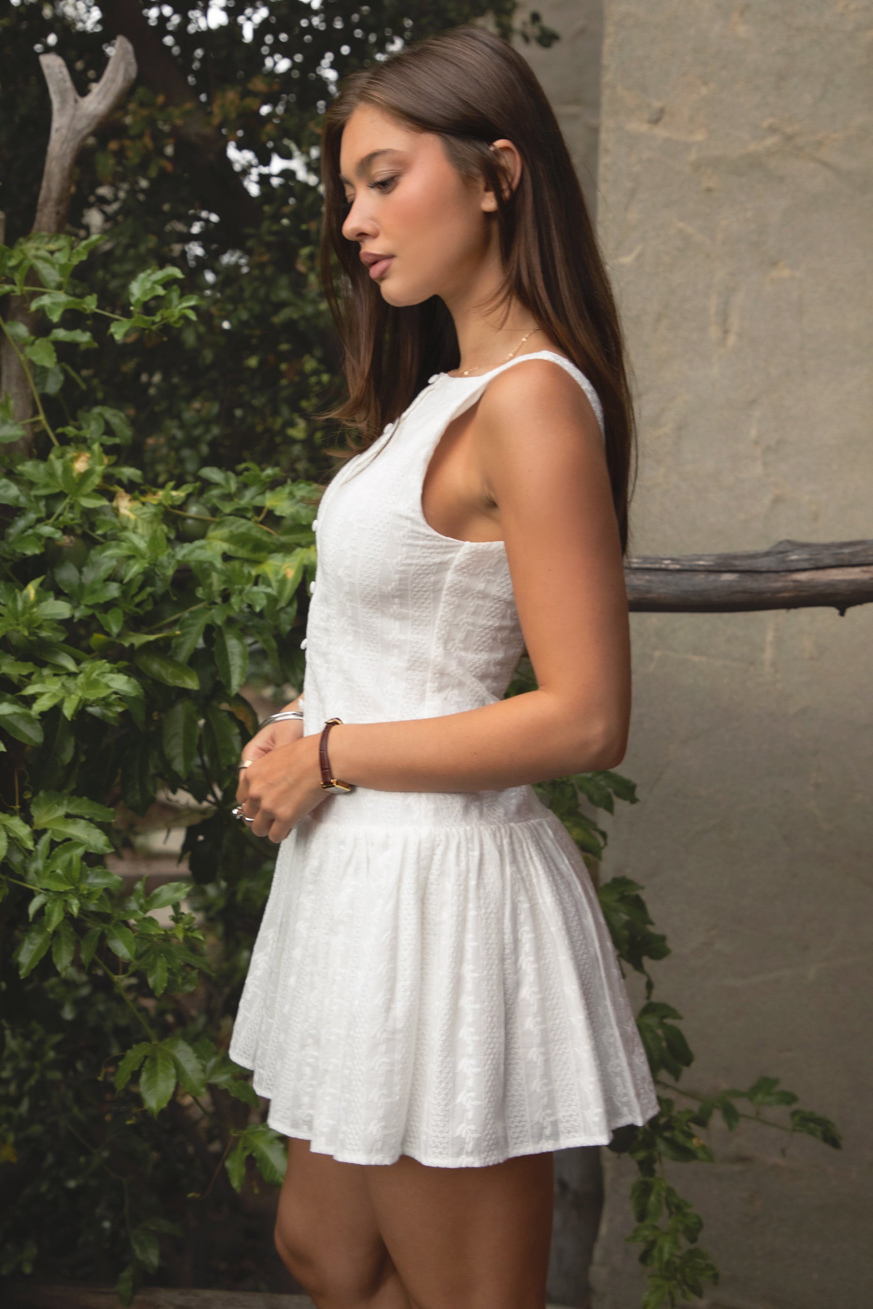 Woman wearing TIC TOC DH31913-AMABEL AMABEL embroidered drop waist mini dress made from allover eyelet embroidered cotton, featuring a clean boat neckline, sleeveless slim fit bodice, faux button front detail, drop waist seam with a flowy mini skirt, open back with zipper closure, and a lightweight fully lined construction, styled for a spring brunch or garden party look.