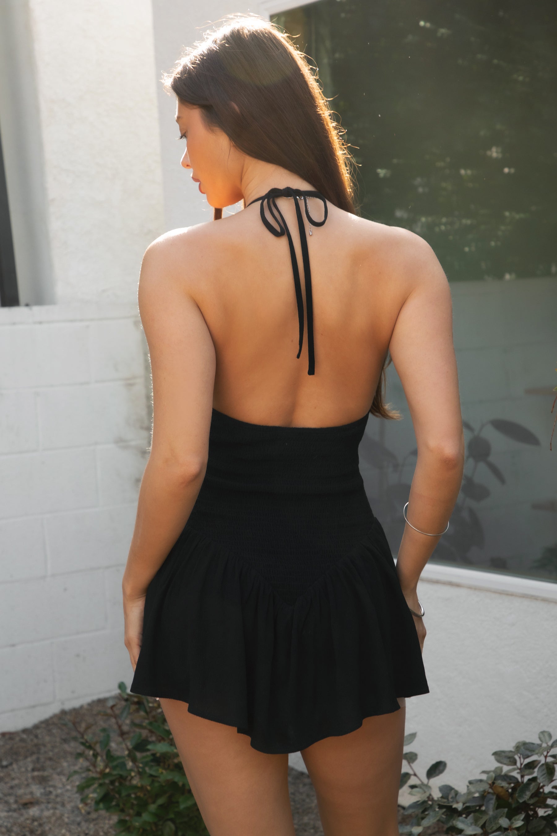 Woman wearing TIC TOC DH31965-CLARISSE CLARISSE crinkle gauze halter mini dress featuring a deep V-neck halter neckline with back tie, open back design, V-shaped fitted bodice with front bow detail, mini length skirt with sheer crinkle gauze overlay and interior lining, and a lightweight flowy silhouette, styled with simple sandals for an airy summer look.