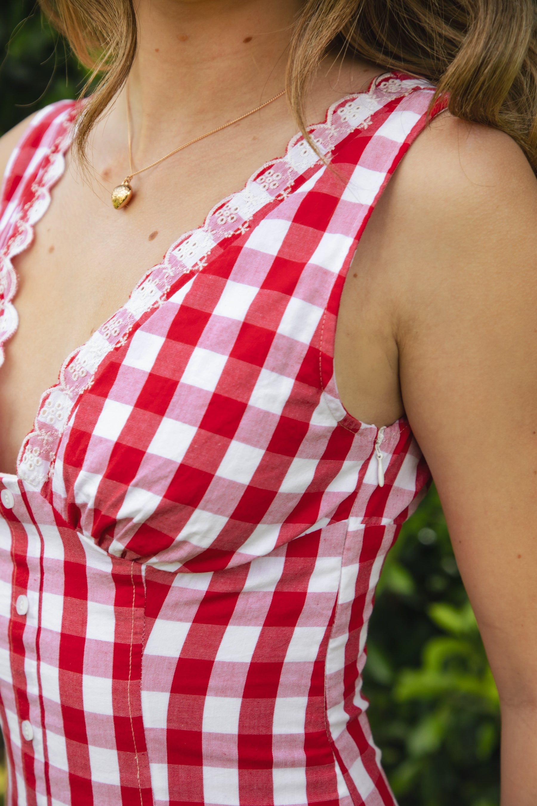 Woman wearing CHERYN V-neck gingham poplin mini dress featuring a classic gingham check pattern, scalloped lace trim neckline, sleeveless fit-and-flare bodice, decorative faux button detail, smocked back panel, hidden side zipper, and floral eyelet lace hem.