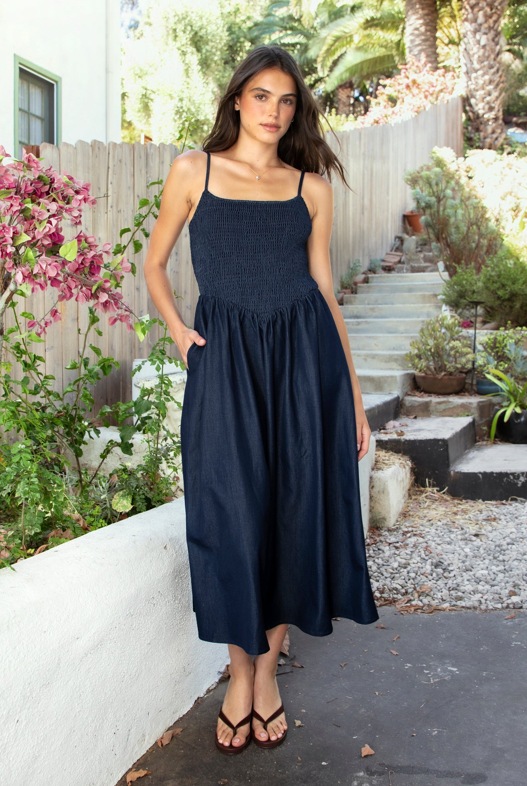 Woman wearing MAYREN smocked bow back denim Tencel maxi dress featuring soft denim Tencel fabric, comfortable stretch, straight neckline, adjustable spaghetti straps, smocked bodice, statement open back with oversized self tie bow detail, side seam pockets, and flowing maxi length skirt.