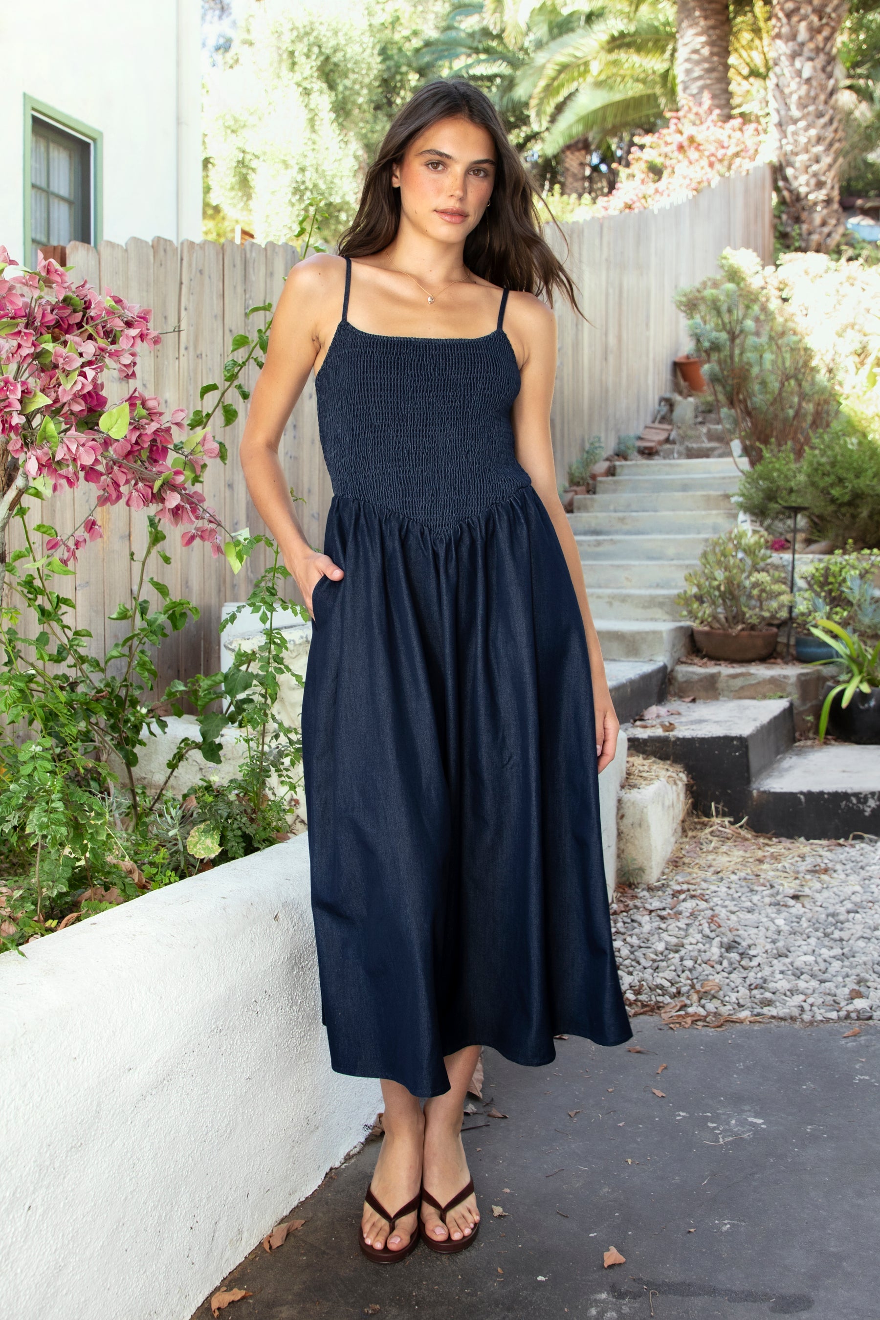 Woman wearing MAYREN smocked bow back denim Tencel maxi dress featuring soft denim Tencel fabric, comfortable stretch, straight neckline, adjustable spaghetti straps, smocked bodice, statement open back with oversized self tie bow detail, side seam pockets, and flowing maxi length skirt.