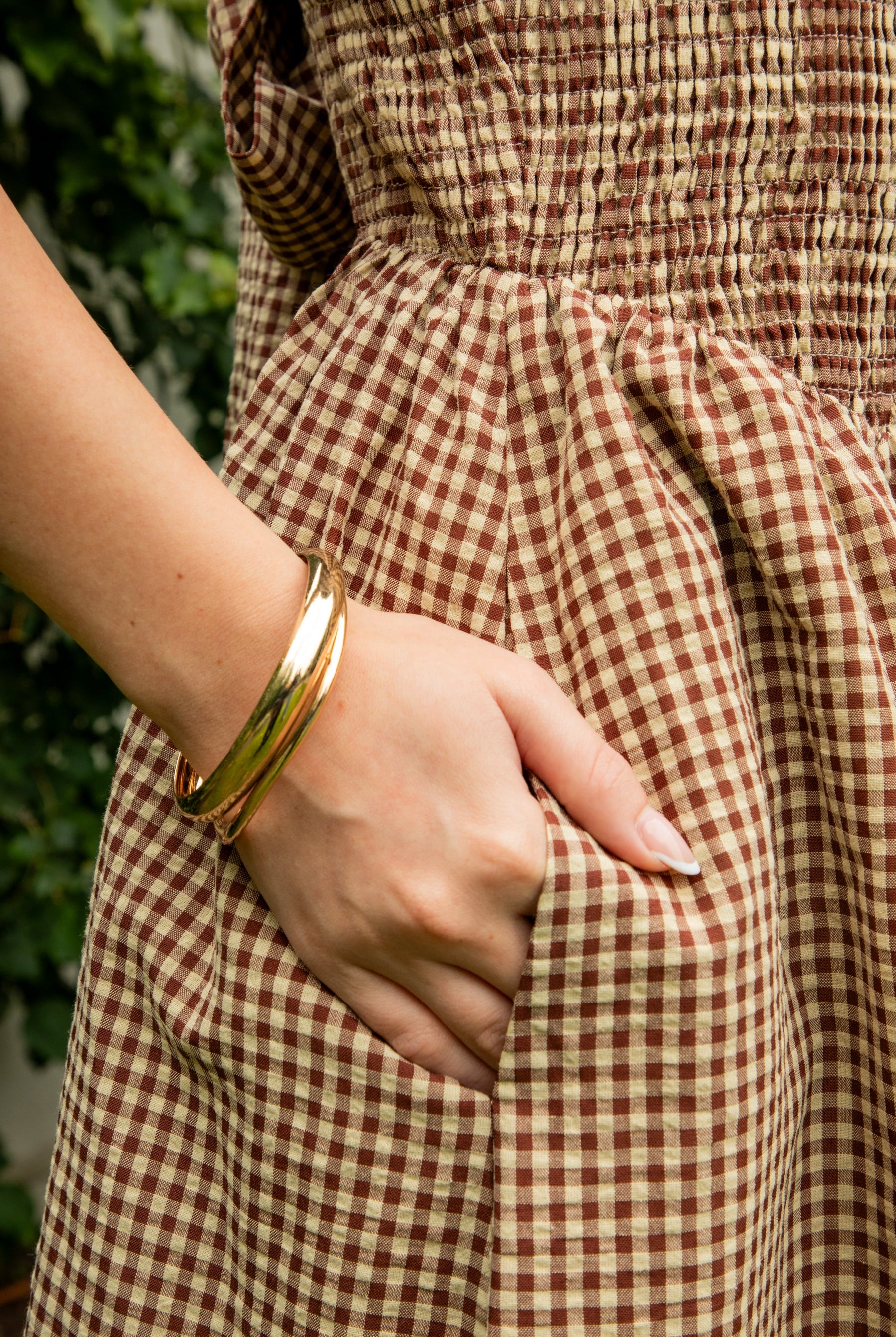 Woman wearing MAYREN gingham smocked midi dress featuring a classic gingham check pattern, fully smocked bodice, adjustable spaghetti straps, square neckline, open back with adjustable tie detail, subtle cutout accent, side seam pockets, and a flowing gathered midi skirt.