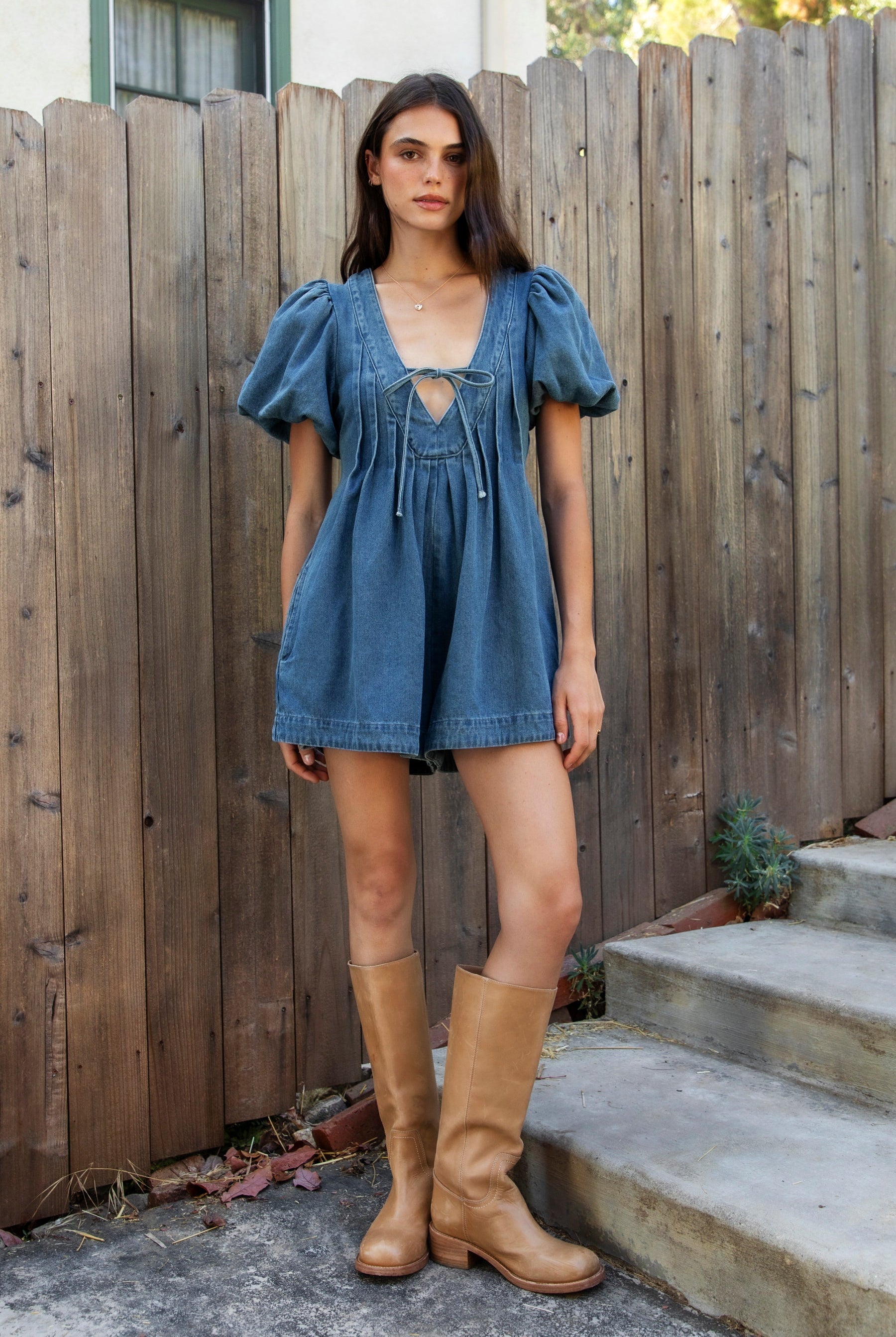 Woman wearing DELANEY soft denim puff sleeve denim romper featuring soft denim fabric, comfortable structured feel, deep V neckline, adjustable front tie detail, statement puff sleeves with gathered volume, pleated bodice, relaxed fit silhouette, and hidden side zipper closure.