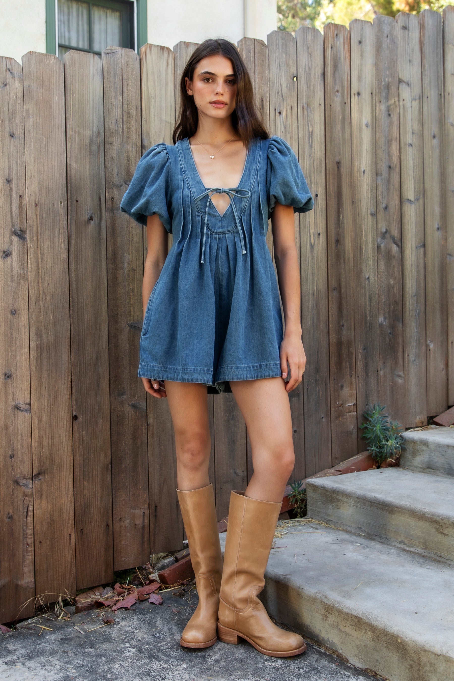 Woman wearing DELANEY soft denim puff sleeve denim romper featuring soft denim fabric, comfortable structured feel, deep V neckline, adjustable front tie detail, statement puff sleeves with gathered volume, pleated bodice, relaxed fit silhouette, and hidden side zipper closure.