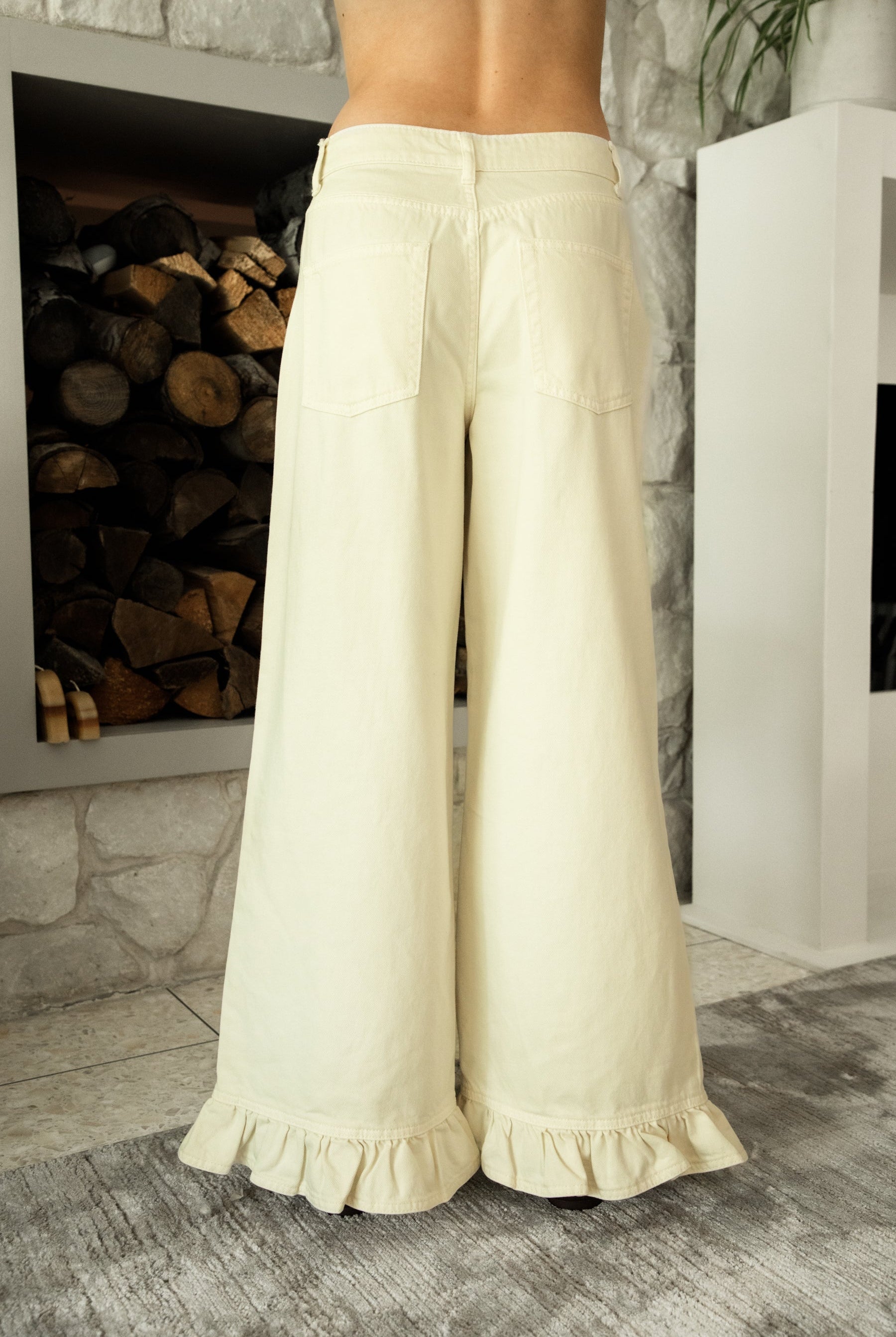 Woman wearing POSIE ruffle hem wide leg pants featuring soft cotton denim, relaxed mid rise fit, classic five pocket design, button and zip closure, and a feminine ruffle hem detail for easy casual styling with tanks, blouses, and cropped knit tops.