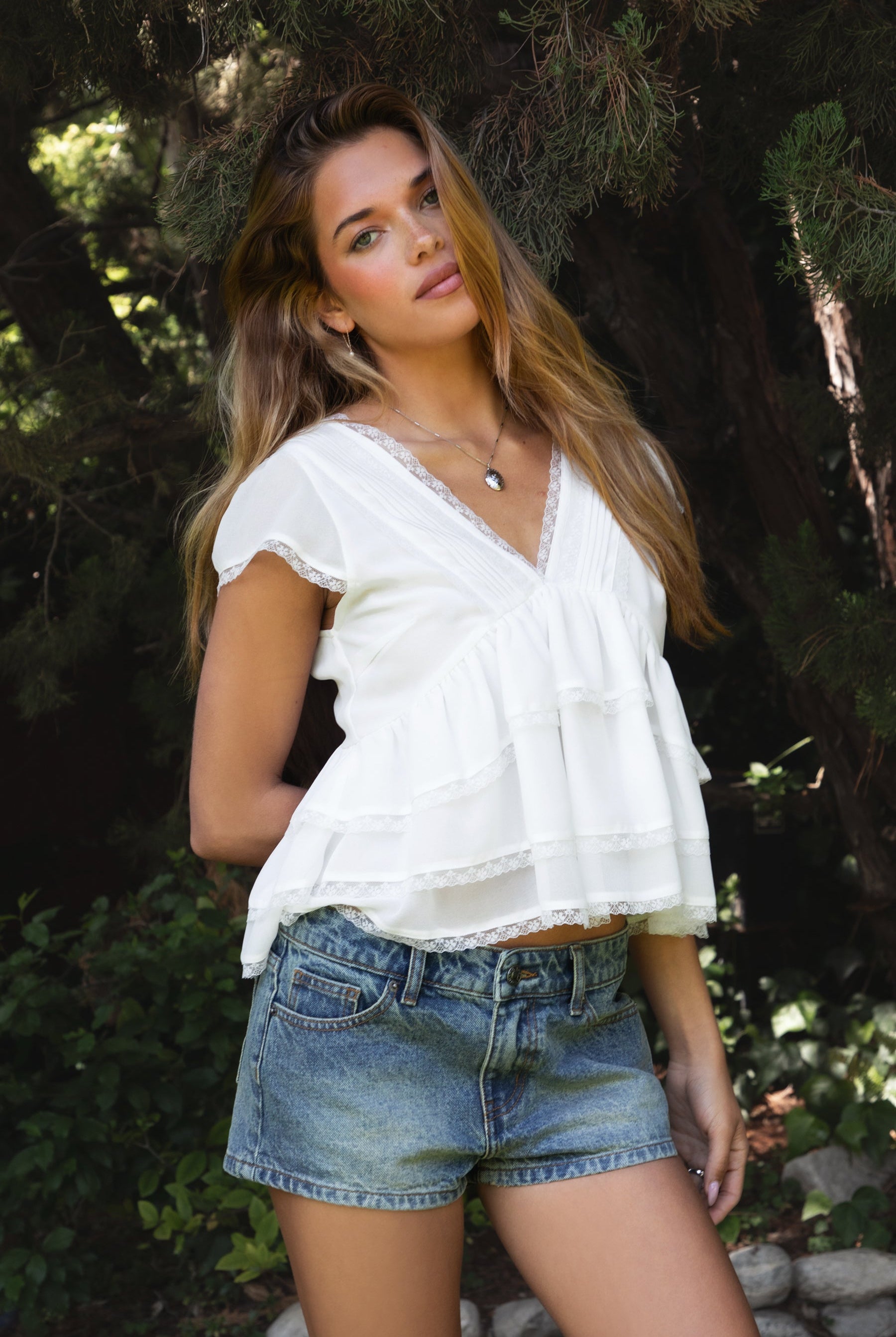 Woman wearing JULIETTE V-neck tiered ruffle short sleeve blouse featuring lightweight airy fabric, pleated V-neckline with lace trim, soft ruffle sleeves, tiered ruffle hem with lace detail, and a V-cut back with adjustable strap for a relaxed flowy silhouette.