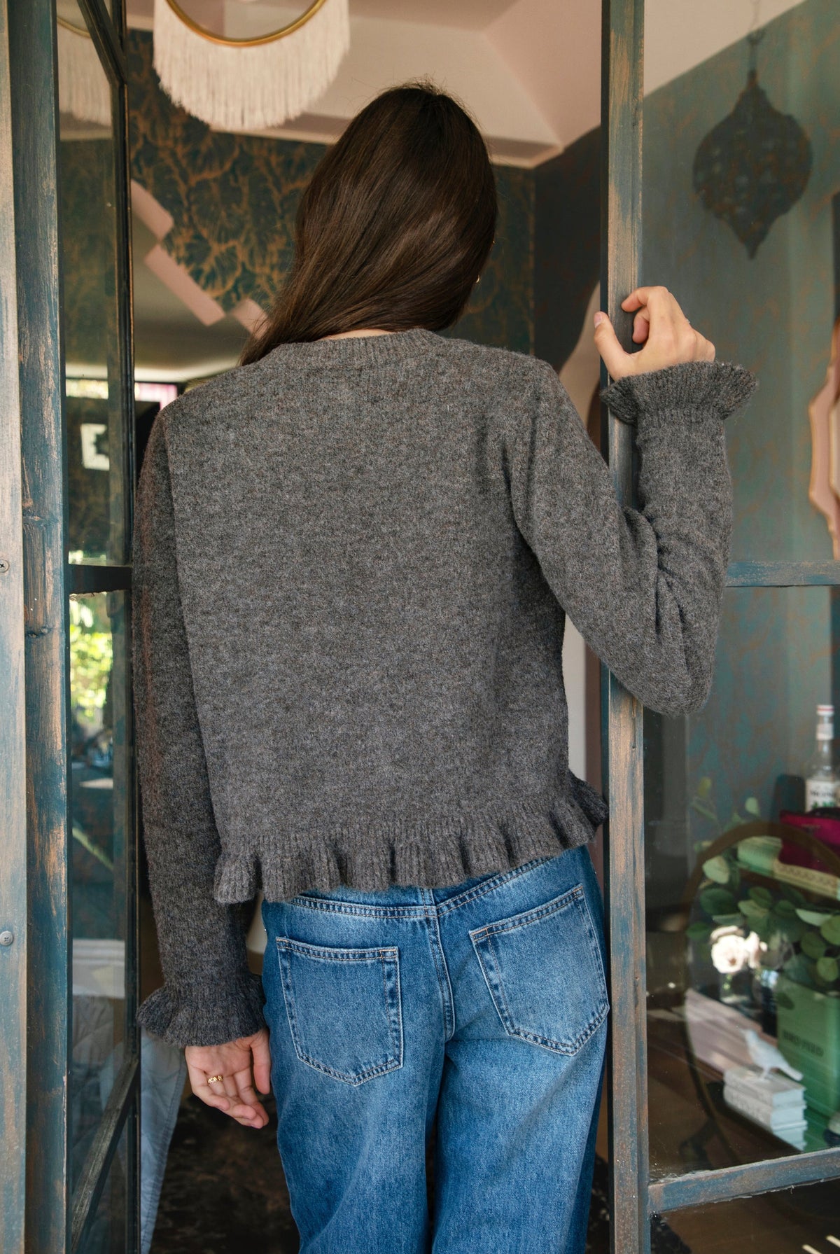 Woman wearing FRILLIA ruffle trim button front knit cardigan featuring soft brushed sweater knit fabric, cozy stretch feel, round neckline, button front closure, delicate ruffle trim along the front placket and hem, long sleeves with flared ruffle cuffs, and relaxed cropped silhouette.