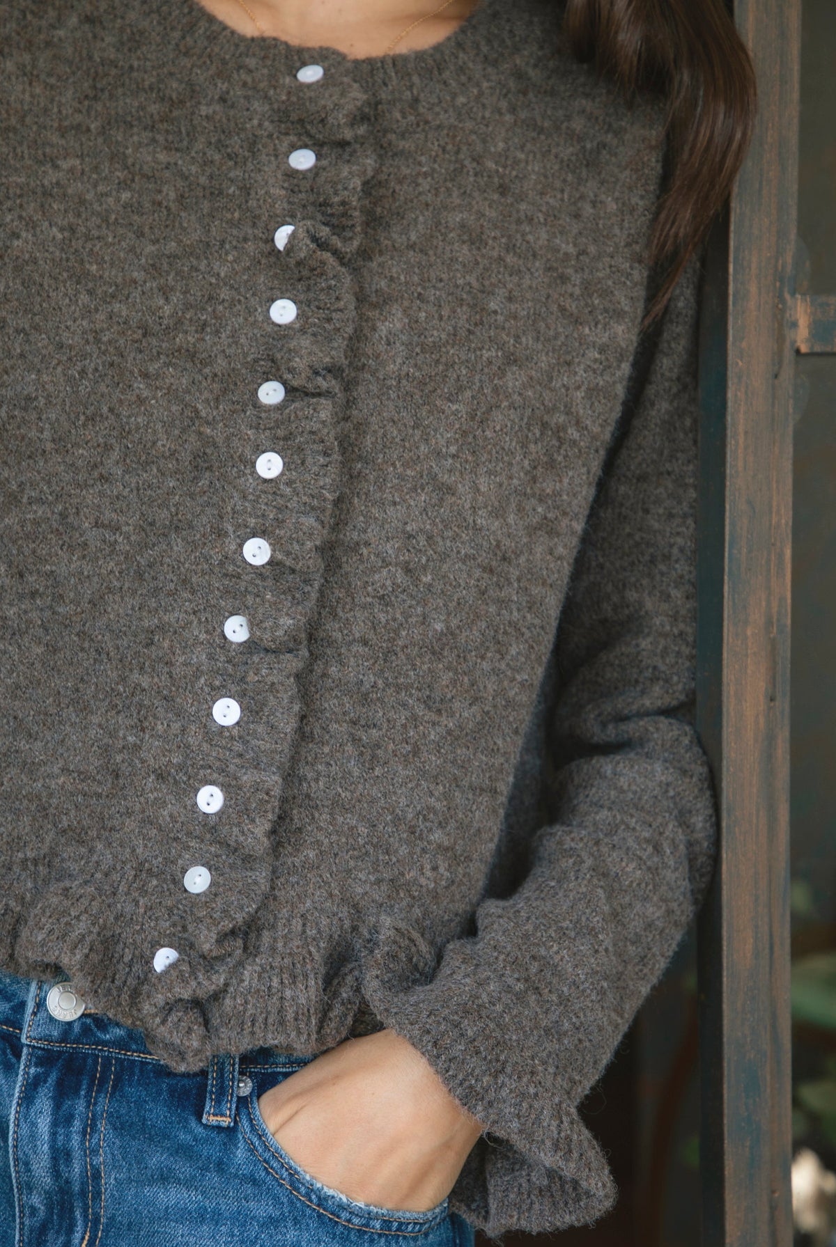 Woman wearing FRILLIA ruffle trim button front knit cardigan featuring soft brushed sweater knit fabric, cozy stretch feel, round neckline, button front closure, delicate ruffle trim along the front placket and hem, long sleeves with flared ruffle cuffs, and relaxed cropped silhouette.