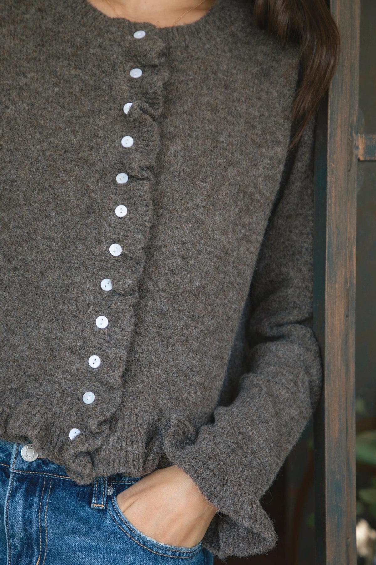 Woman wearing FRILLIA ruffle trim button front knit cardigan featuring soft brushed sweater knit fabric, cozy stretch feel, round neckline, button front closure, delicate ruffle trim along the front placket and hem, long sleeves with flared ruffle cuffs, and relaxed cropped silhouette.
