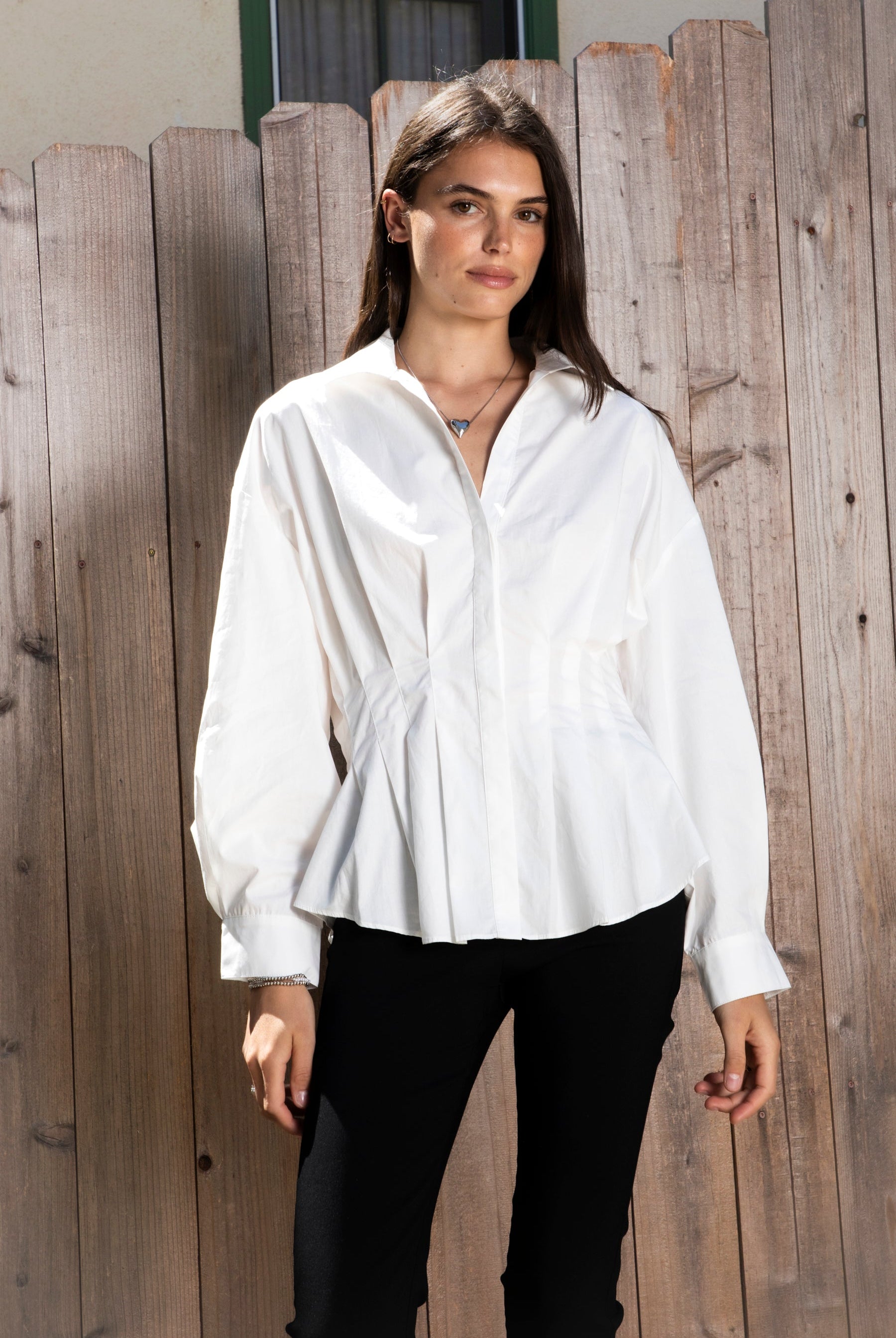 Woman wearing VIENNA tailored poplin peplum puff sleeve shirt featuring crisp cotton poplin fabric, structured feel, classic collared V neckline, hidden button front closure, defined waist seaming, soft pleated peplum hem, relaxed long balloon sleeves, and button cuffs for elevated everyday styling.