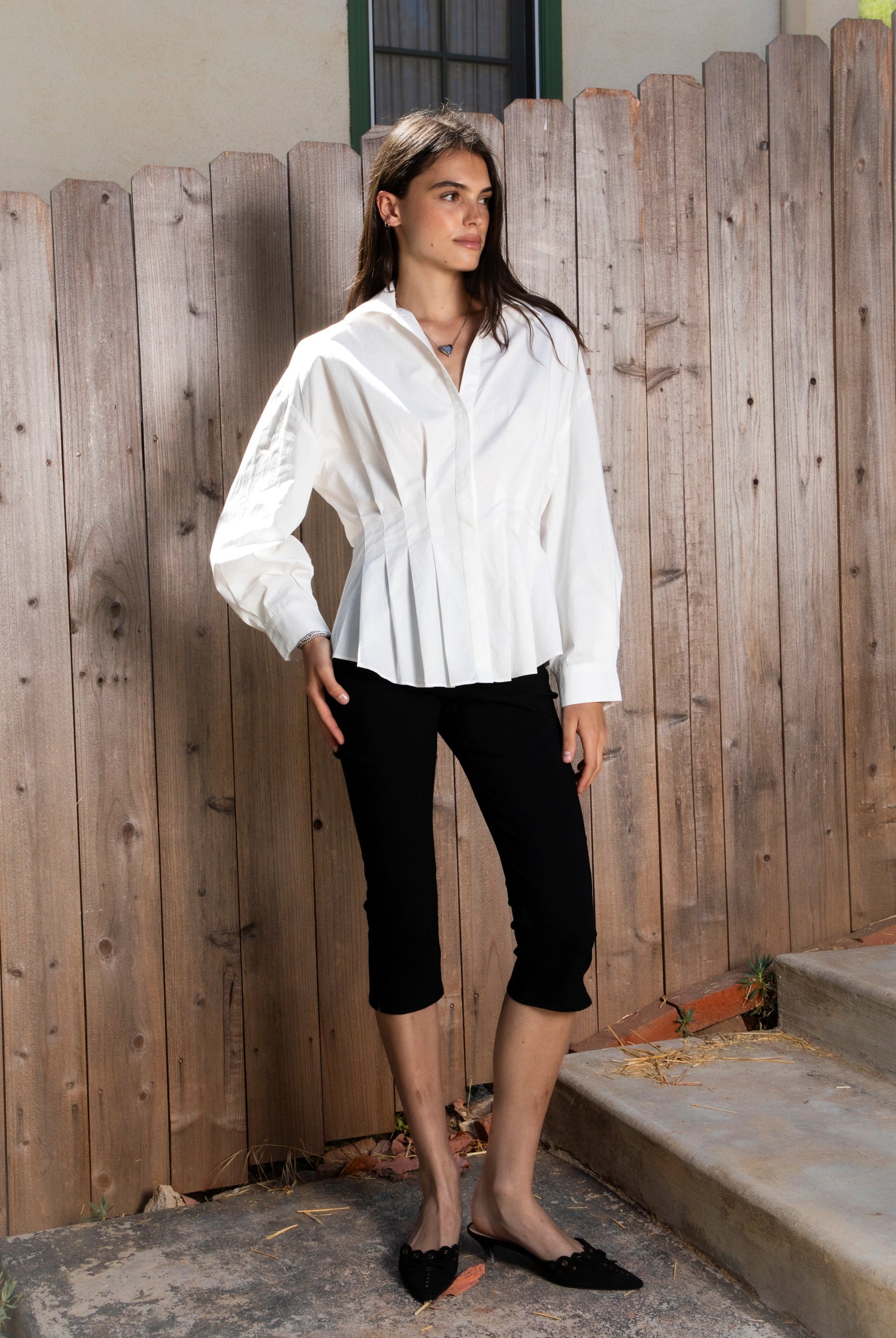 Woman wearing VIENNA tailored poplin peplum puff sleeve shirt featuring crisp cotton poplin fabric, structured feel, classic collared V neckline, hidden button front closure, defined waist seaming, soft pleated peplum hem, relaxed long balloon sleeves, and button cuffs for elevated everyday styling.
