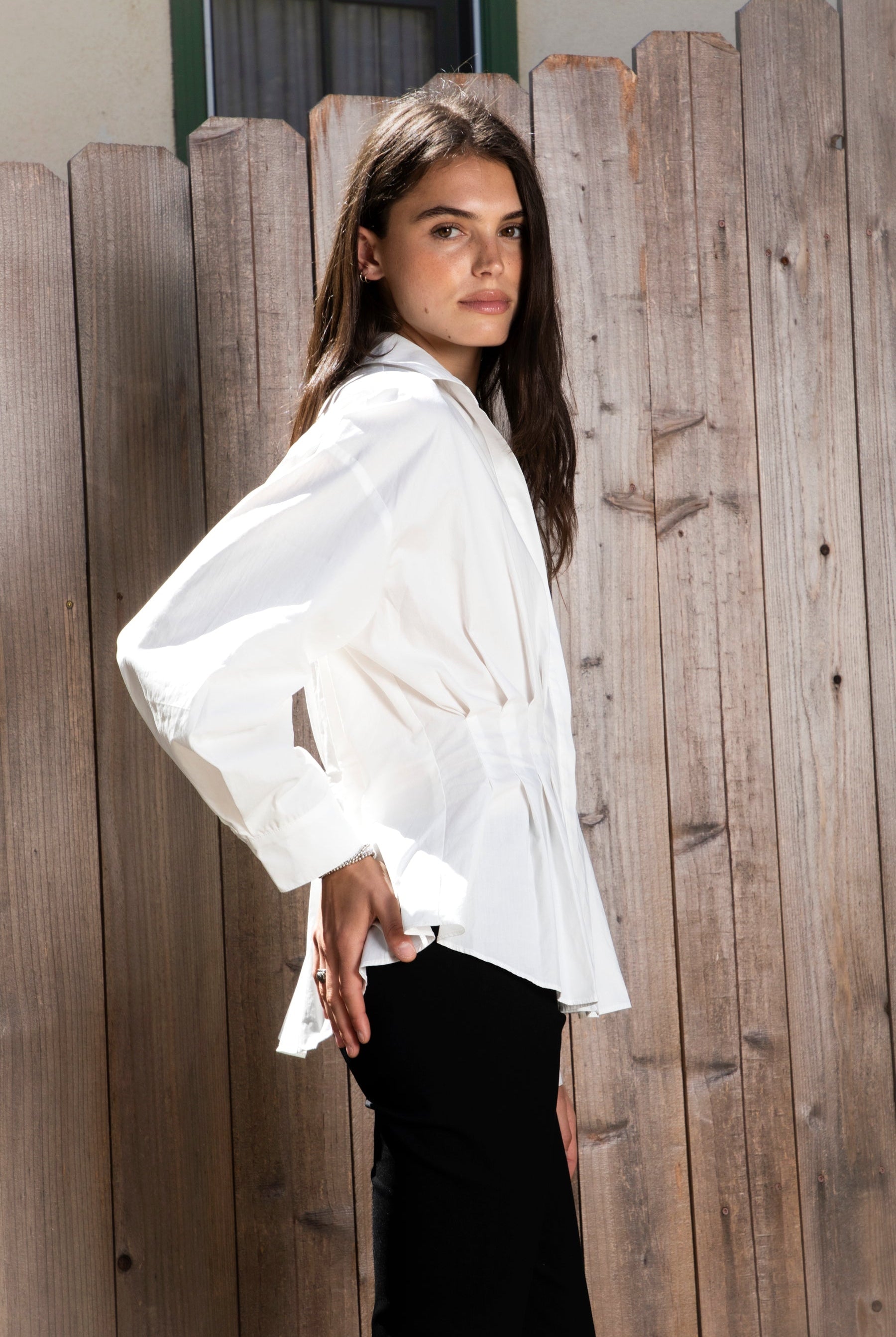 Woman wearing VIENNA tailored poplin peplum puff sleeve shirt featuring crisp cotton poplin fabric, structured feel, classic collared V neckline, hidden button front closure, defined waist seaming, soft pleated peplum hem, relaxed long balloon sleeves, and button cuffs for elevated everyday styling.