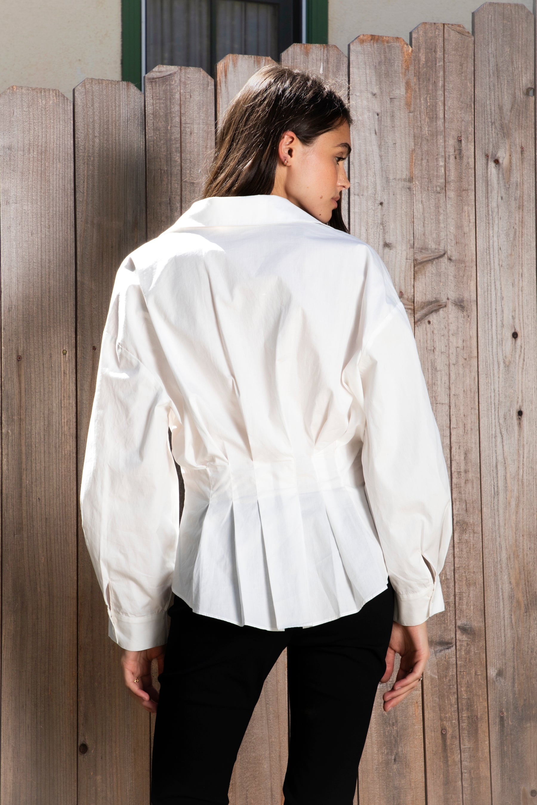 Woman wearing VIENNA tailored poplin peplum puff sleeve shirt featuring crisp cotton poplin fabric, structured feel, classic collared V neckline, hidden button front closure, defined waist seaming, soft pleated peplum hem, relaxed long balloon sleeves, and button cuffs for elevated everyday styling.