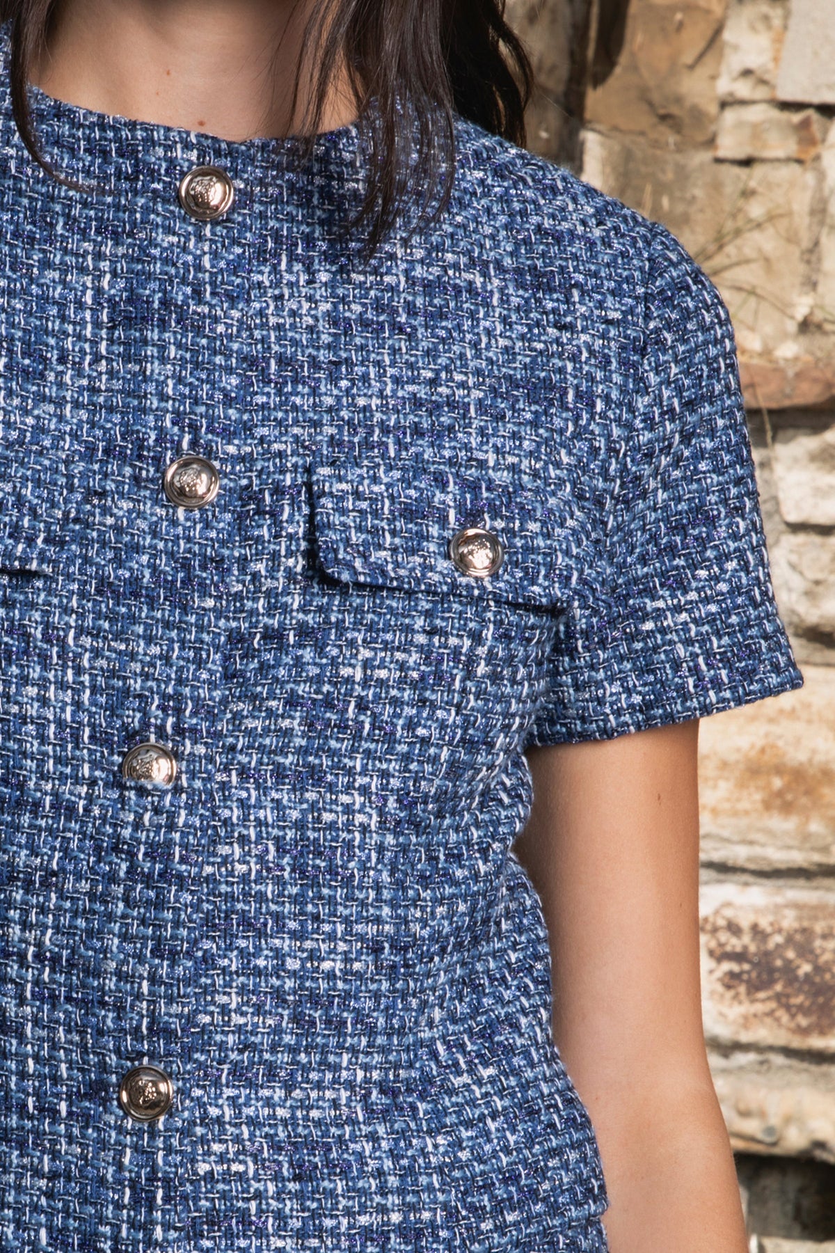 Woman wearing COCO navy blue lurex tweed short sleeve top featuring shimmering textured tweed fabric, subtle metallic lurex detail, round neckline, fitted structured bodice, cropped length, elegant gold point buttons, decorative faux chest pockets, and fully lined construction designed to coordinate with the matching COCO skort.