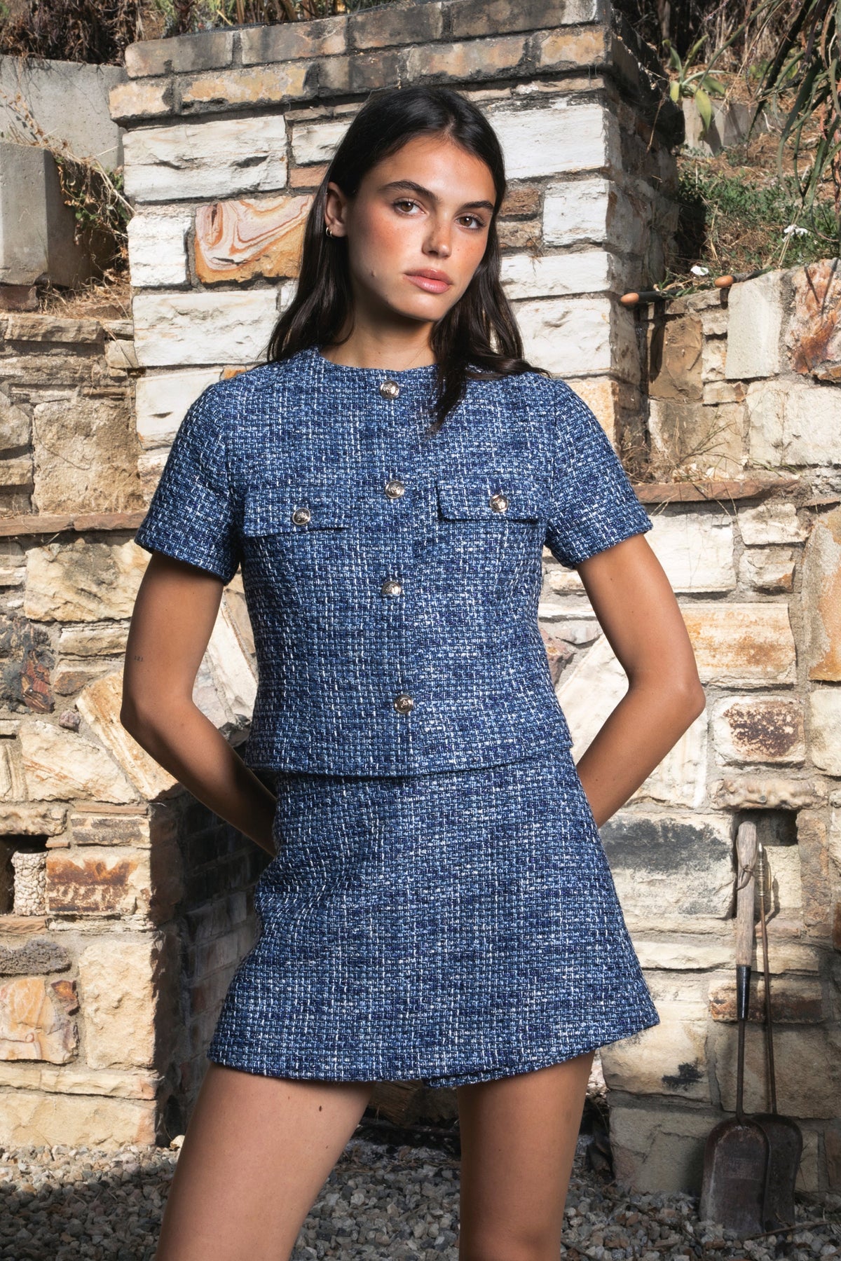 Woman wearing COCO navy blue lurex tweed short sleeve top featuring shimmering textured tweed fabric, subtle metallic lurex detail, round neckline, fitted structured bodice, cropped length, elegant gold point buttons, decorative faux chest pockets, and fully lined construction designed to coordinate with the matching COCO skort.
