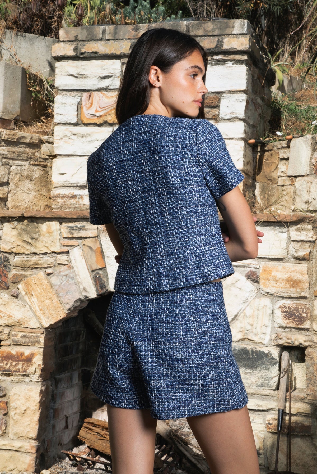 Woman wearing COCO navy blue lurex tweed short sleeve top featuring shimmering textured tweed fabric, subtle metallic lurex detail, round neckline, fitted structured bodice, cropped length, elegant gold point buttons, decorative faux chest pockets, and fully lined construction designed to coordinate with the matching COCO skort.