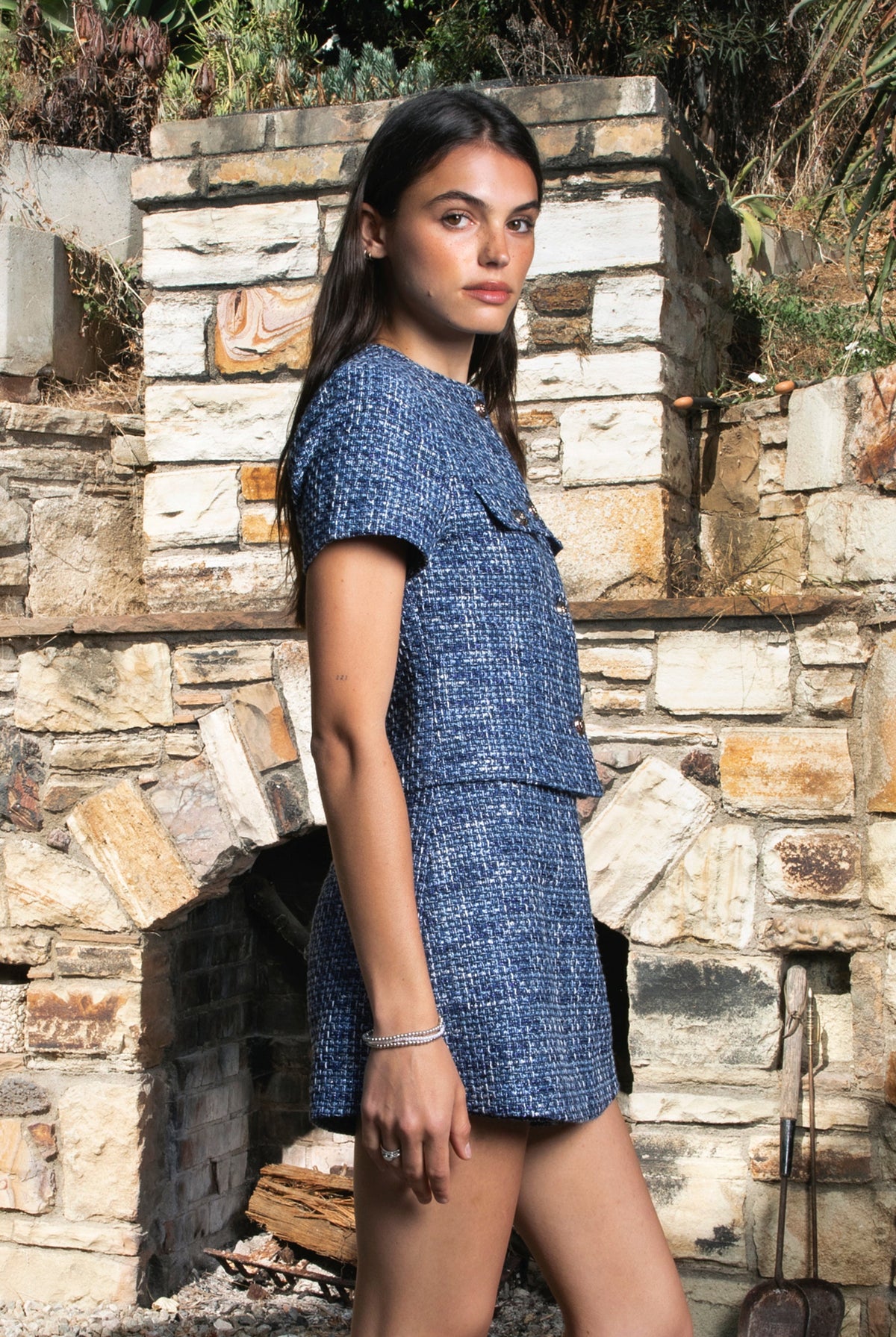 Woman wearing COCO navy blue lurex tweed short sleeve top featuring shimmering textured tweed fabric, subtle metallic lurex detail, round neckline, fitted structured bodice, cropped length, elegant gold point buttons, decorative faux chest pockets, and fully lined construction designed to coordinate with the matching COCO skort.