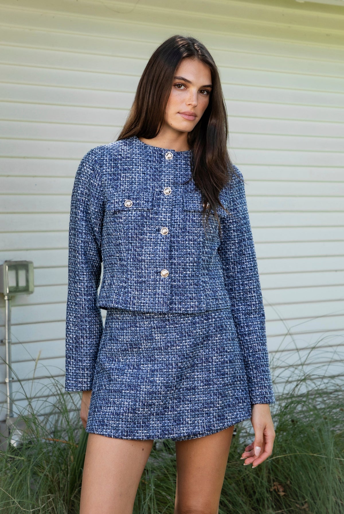 Woman wearing COCO navy blue lurex tweed long sleeve jacket featuring textured lurex tweed fabric, structured cropped silhouette, round neckline, clean collarless design, gold tone statement button front closure, decorative chest flap pockets with matching gold tone buttons, subtle metallic threading, and full lining for a smooth finish.