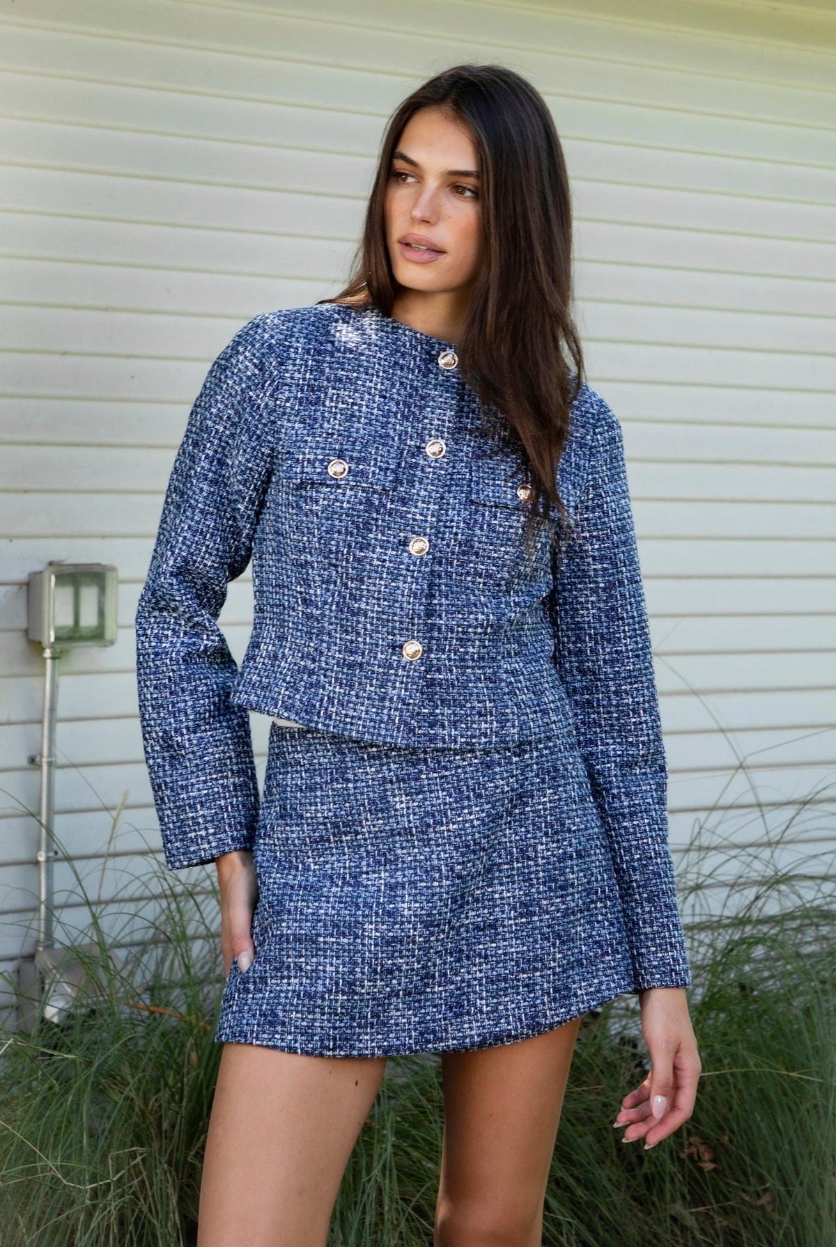 Woman wearing COCO navy blue lurex tweed long sleeve jacket featuring textured lurex tweed fabric, structured cropped silhouette, round neckline, clean collarless design, gold tone statement button front closure, decorative chest flap pockets with matching gold tone buttons, subtle metallic threading, and full lining for a smooth finish.