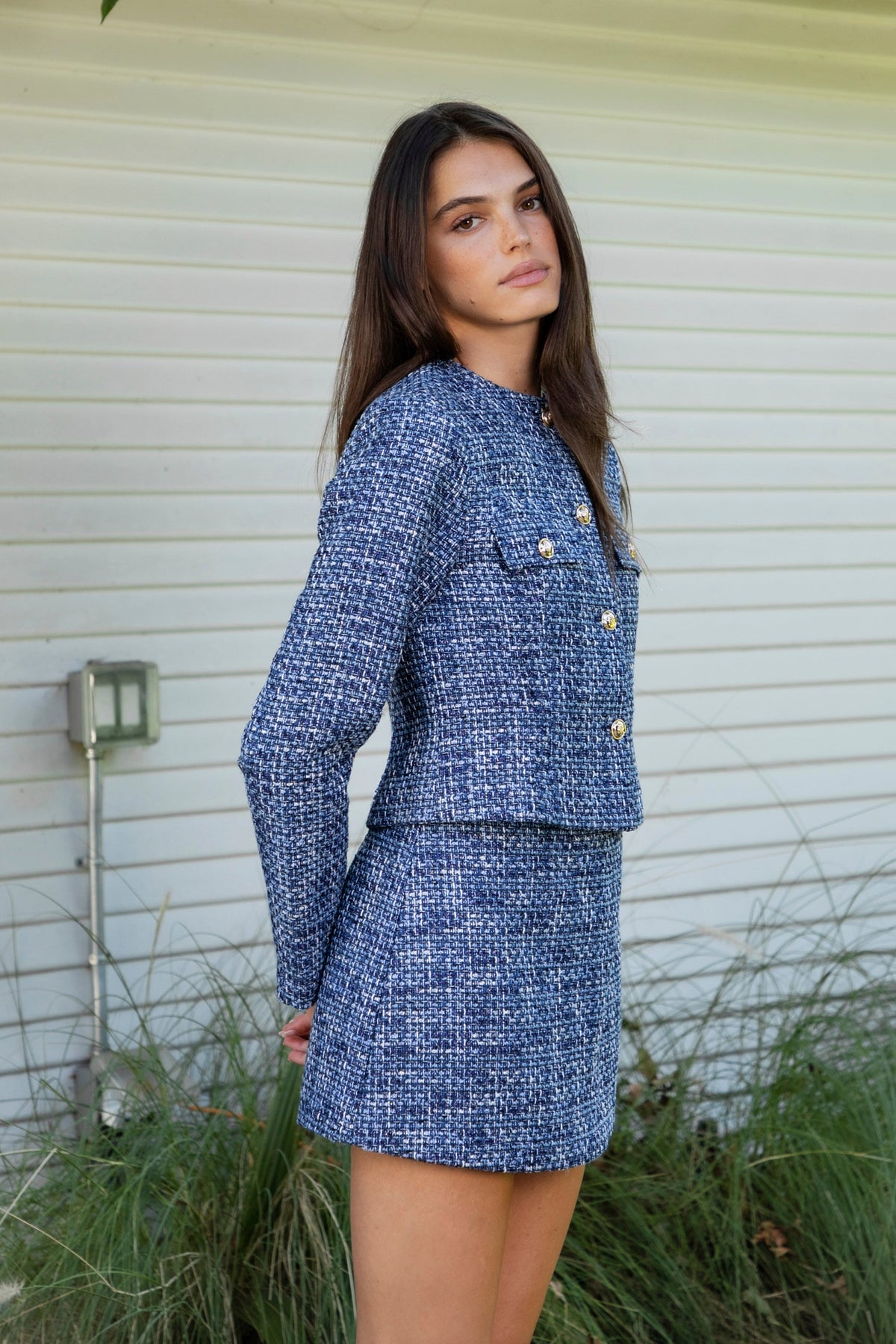 Woman wearing COCO navy blue lurex tweed long sleeve jacket featuring textured lurex tweed fabric, structured cropped silhouette, round neckline, clean collarless design, gold tone statement button front closure, decorative chest flap pockets with matching gold tone buttons, subtle metallic threading, and full lining for a smooth finish.