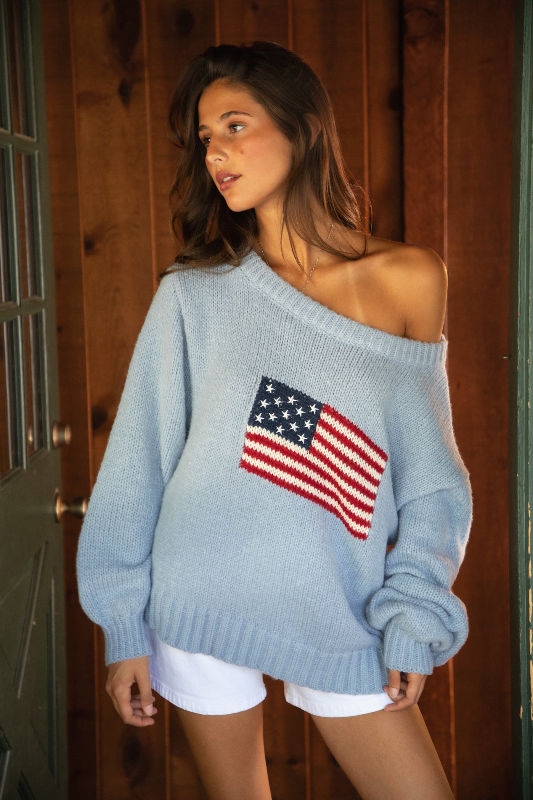 Woman wearing a TIC TOC BRANDI Ribbed Knit Sweater in light blue with off-the-shoulder boat neck, American flag pattern, and semi-oversized fit with white shorts and ugg boots, standing in a wooden cabin.