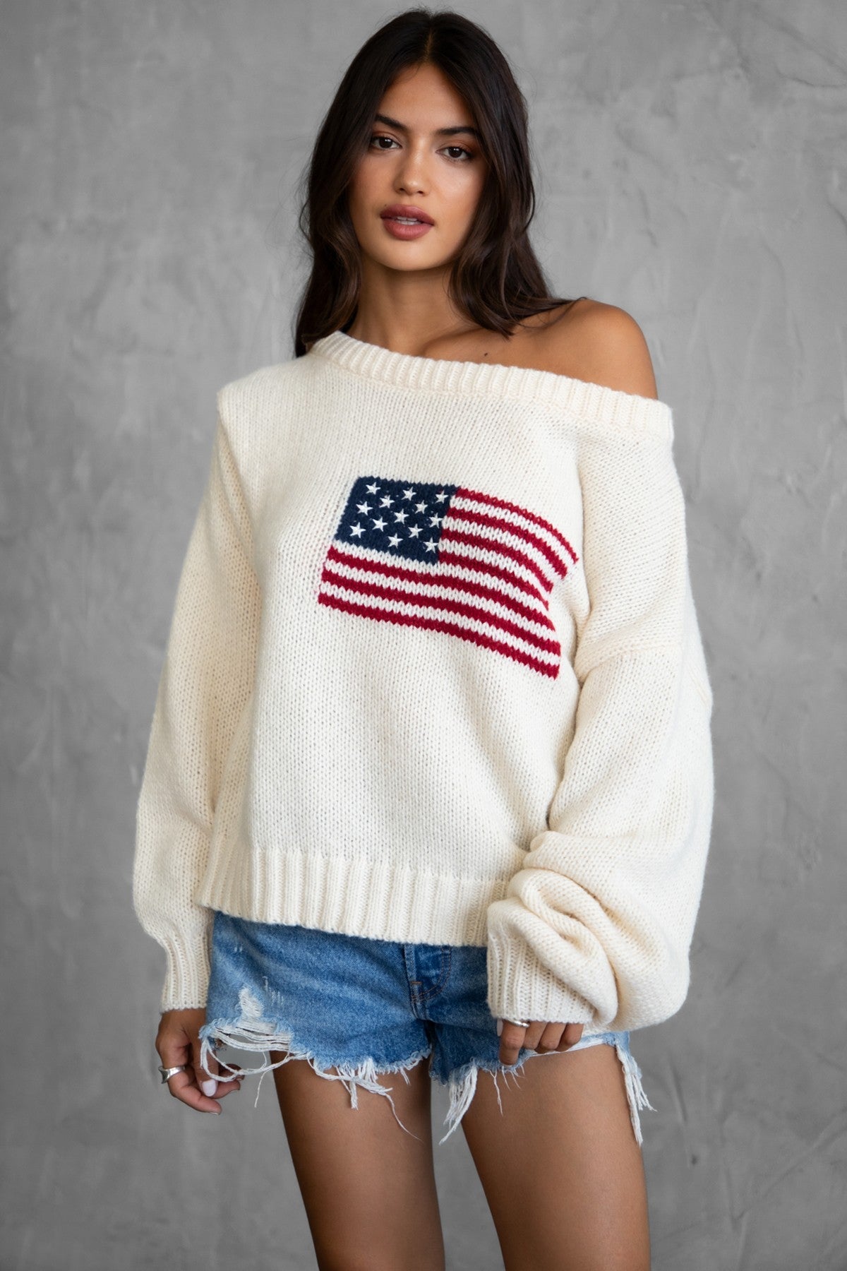 Woman wearing a TIC TOC BRANDI Ribbed Knit Sweater in cream with off-the-shoulder boat neck, American flag pattern, and semi-oversized fit, paired with denim blue shorts.