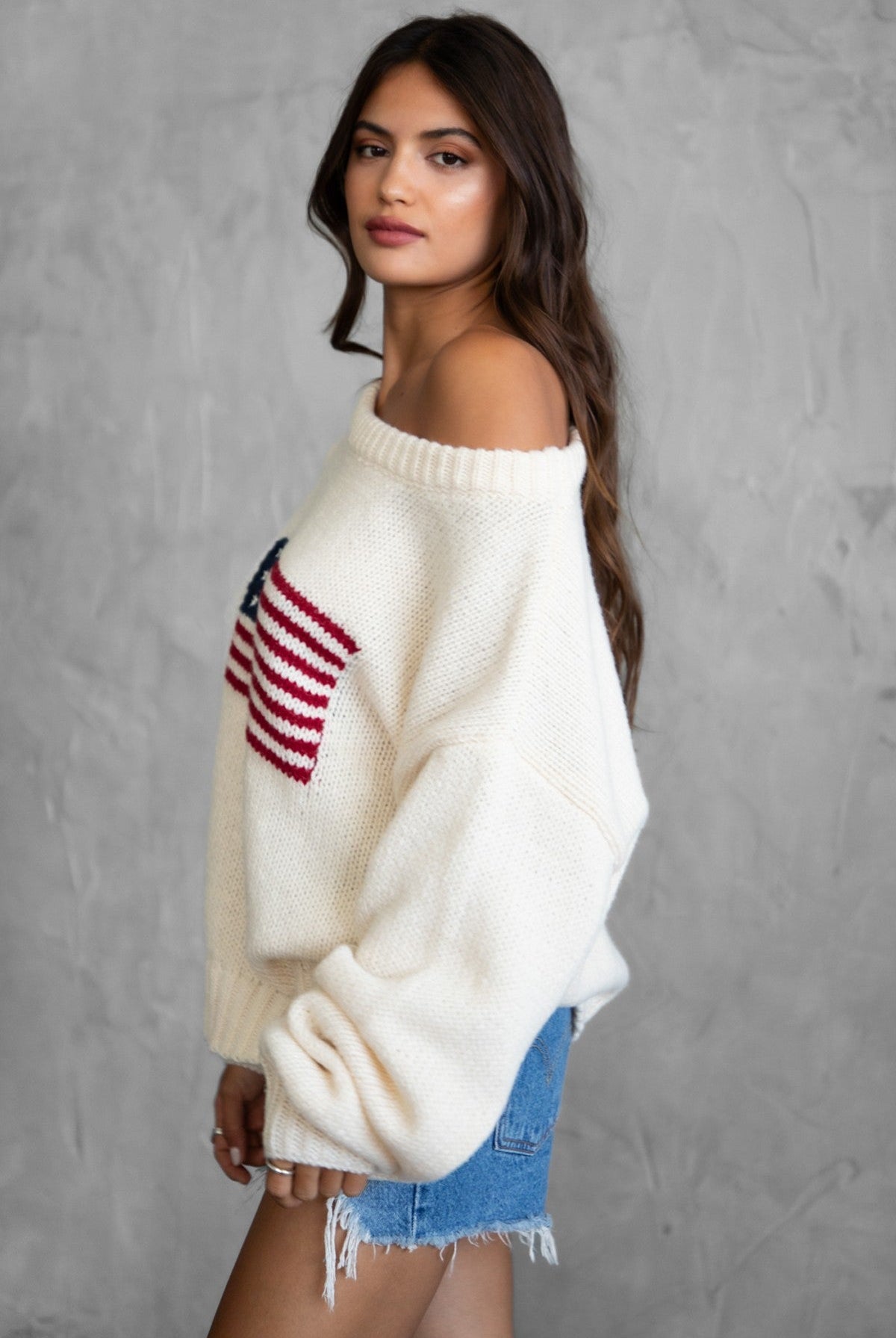 Woman wearing a TIC TOC BRANDI Ribbed Knit Sweater in cream with off-the-shoulder boat neck, American flag pattern, and semi-oversized fit, paired with denim blue shorts.