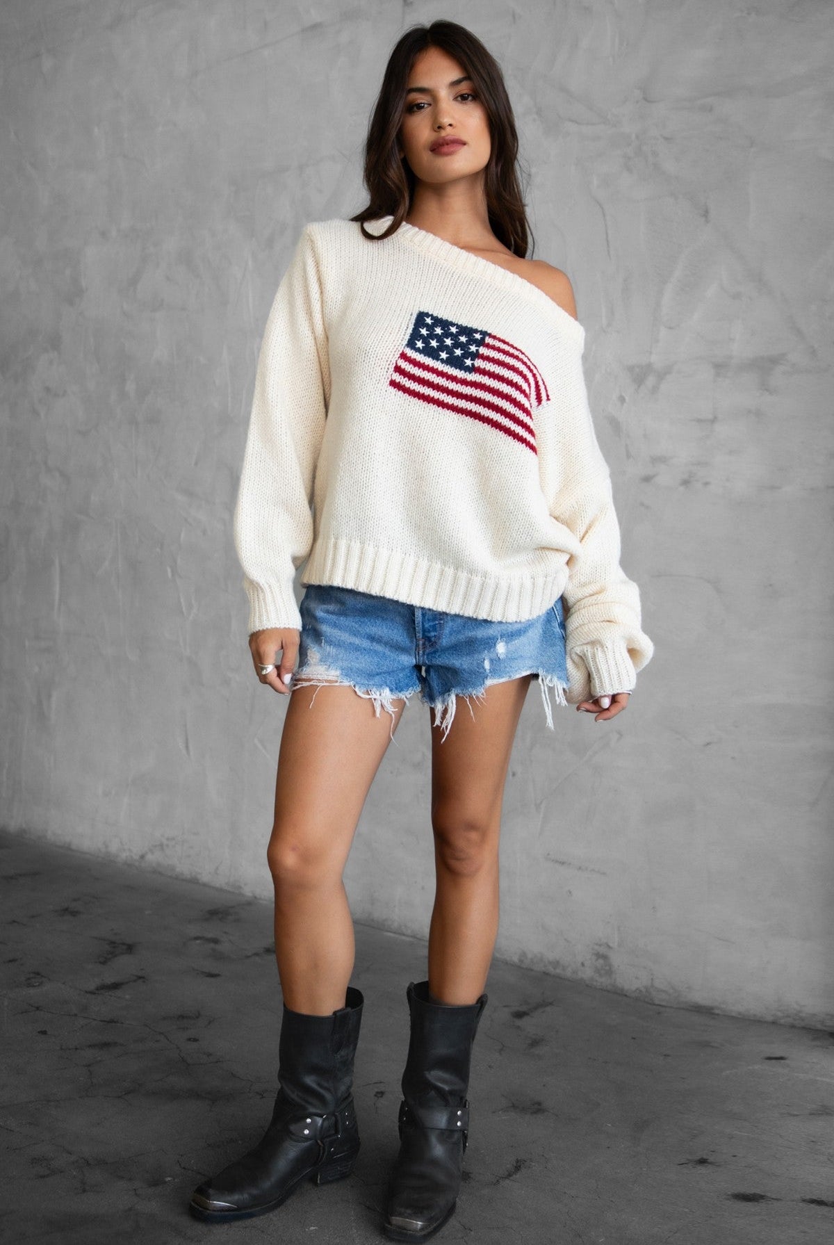 Woman wearing a TIC TOC BRANDI Ribbed Knit Sweater in cream with off-the-shoulder boat neck, American flag pattern, and semi-oversized fit, paired with denim blue shorts.