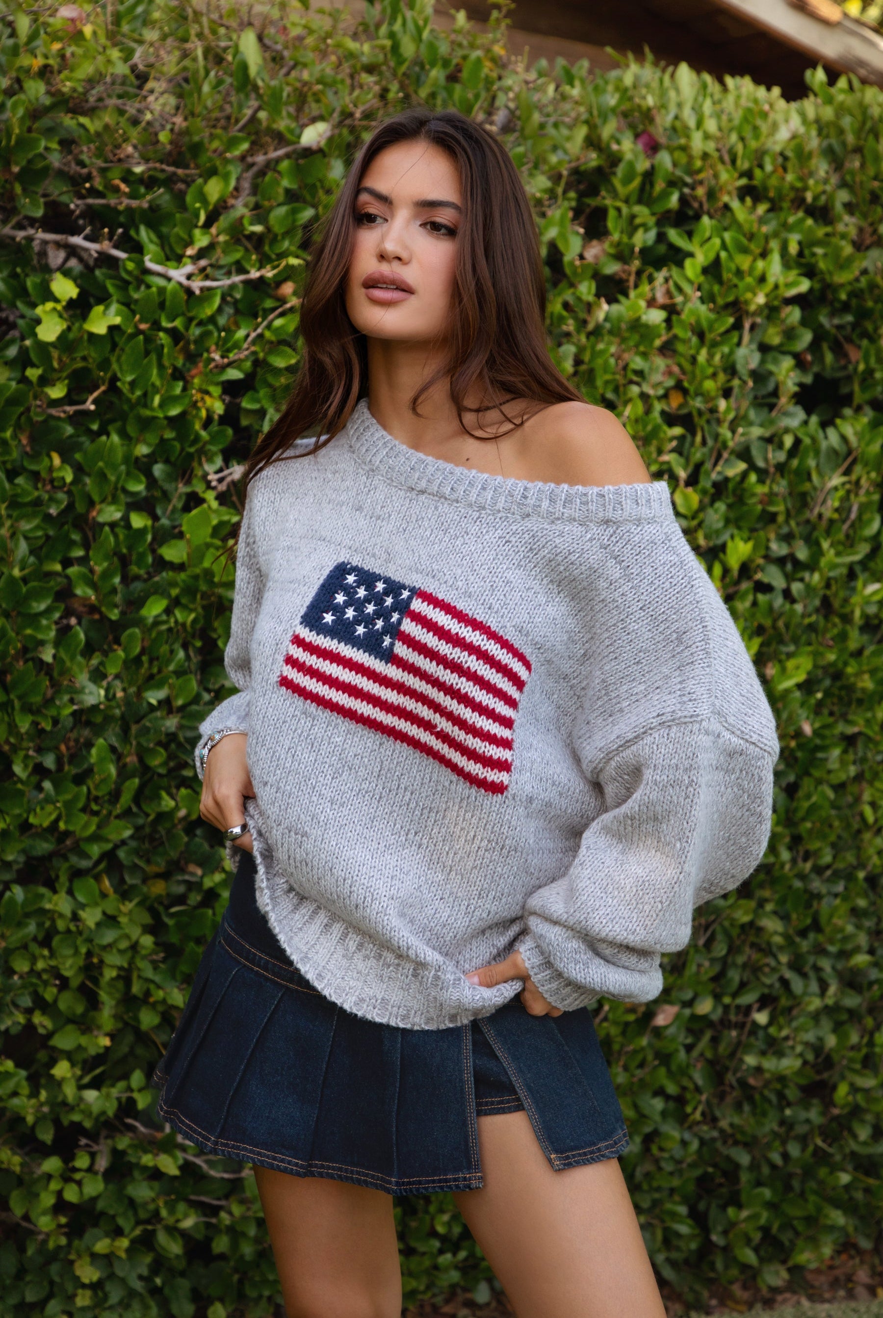 Woman wearing a TIC TOC BRANDI Ribbed Knit Sweater in grey with off-the-shoulder boat neck, American flag pattern, and semi-oversized fit, paired with denim blue skort in front of green bushes.