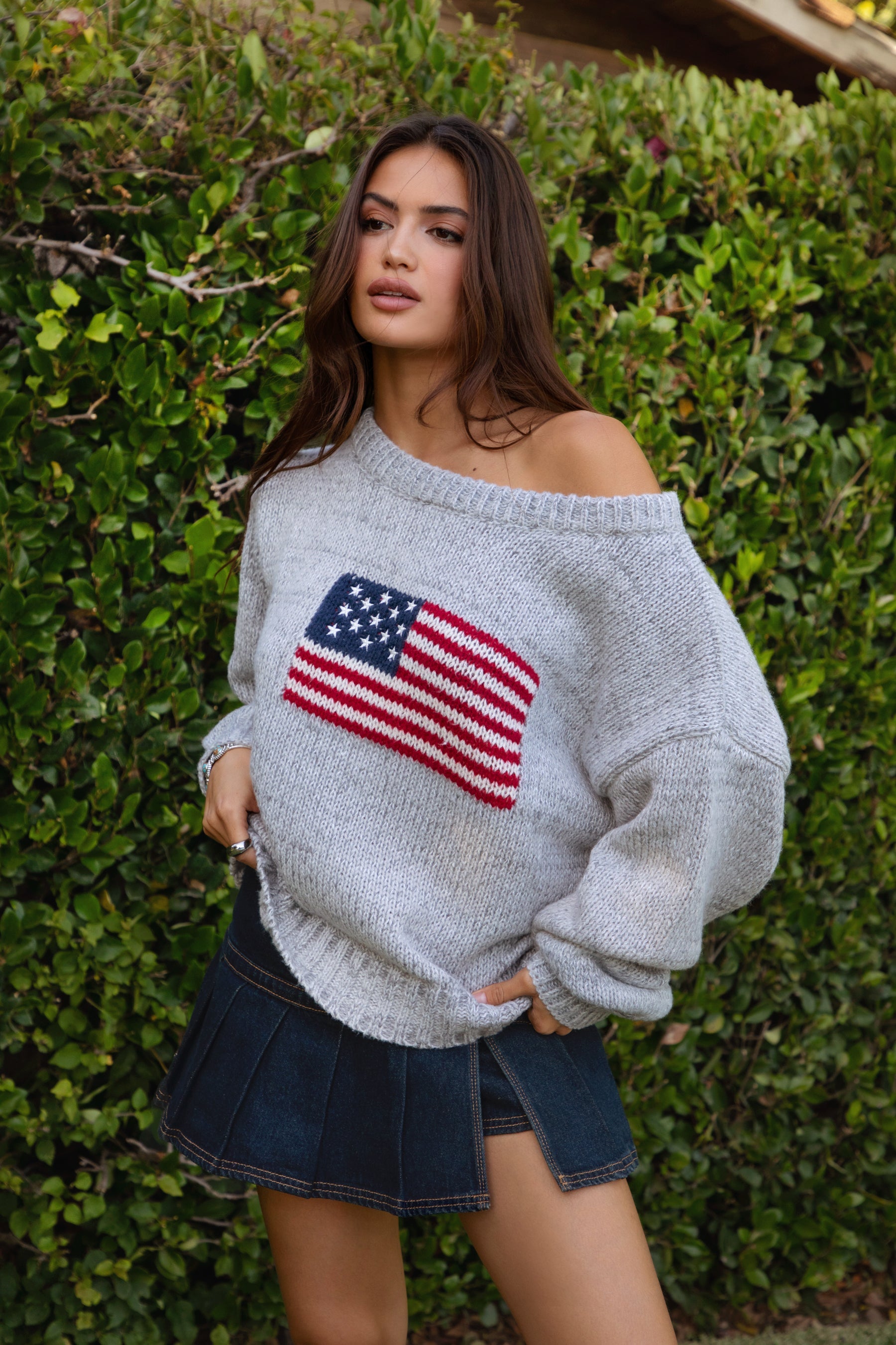Woman wearing a TIC TOC BRANDI Ribbed Knit Sweater in grey with off-the-shoulder boat neck, American flag pattern, and semi-oversized fit, paired with denim blue skort in front of green bushes.