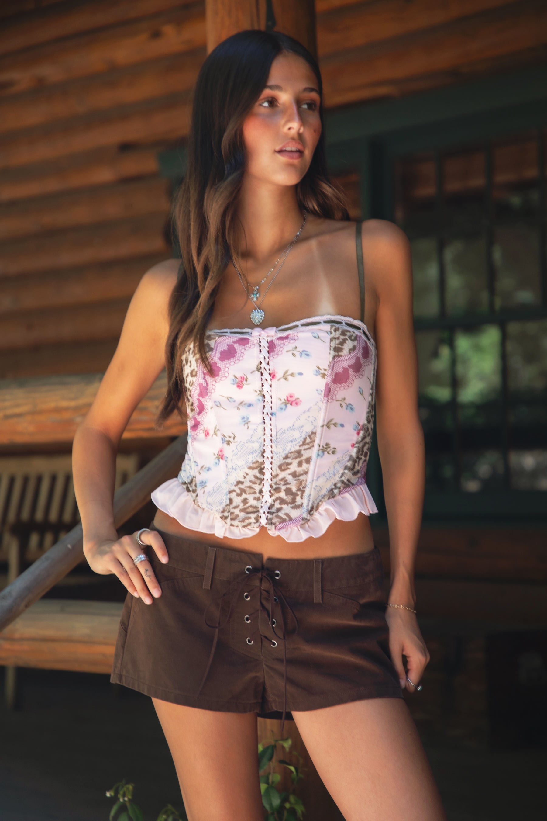 Woman wearing TIC TOC ESME Vintage Inspired Mesh Bustier Corset Top in multi pink featuring lace trim, satin bow, and lace-up front with ruffled details paired with lace up suede shorts in an outdoor cabin setting