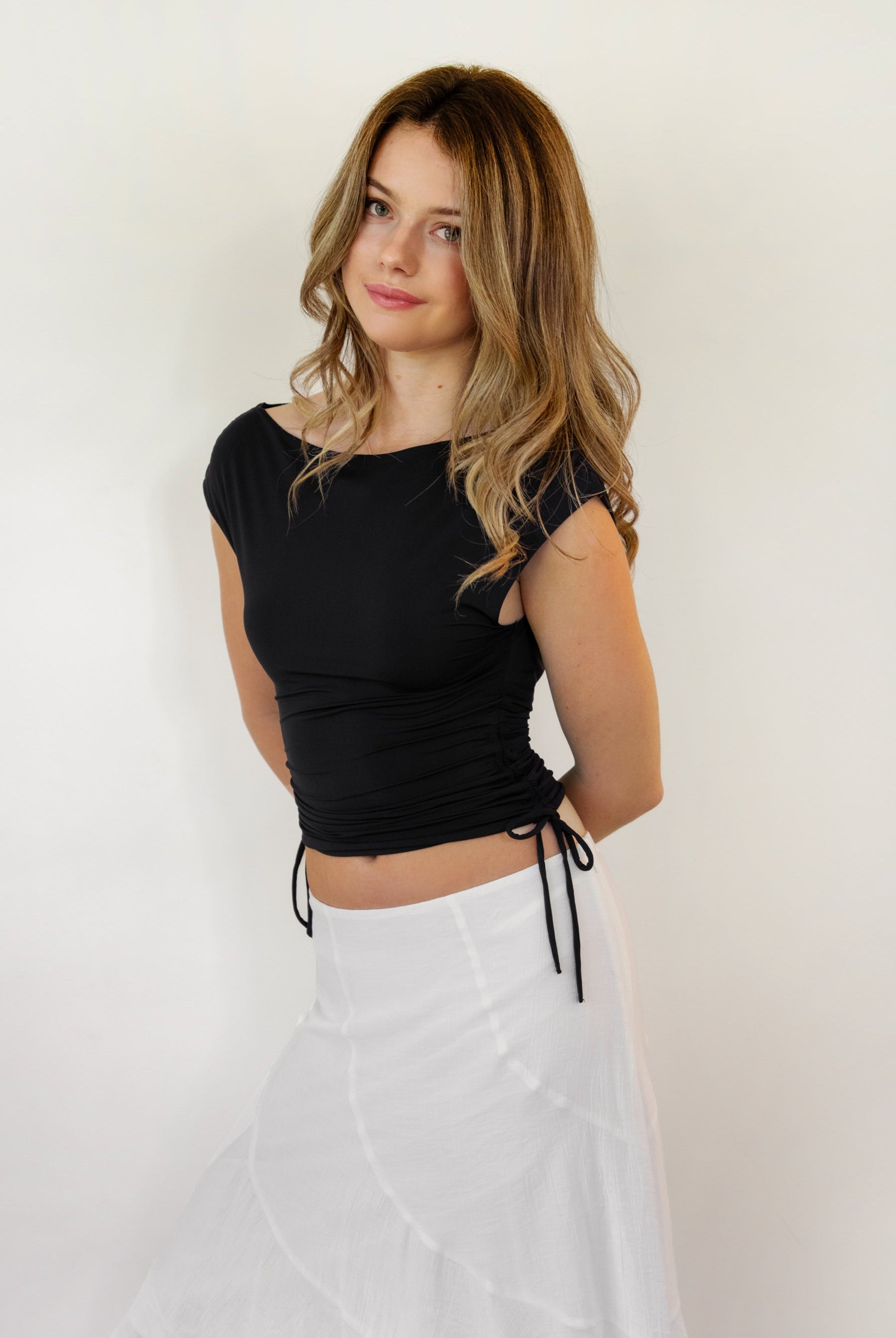 Woman wearing SELENE boat neck cowl back side tie crop top featuring an elegant boat neckline, draped cowl back, adjustable back strap, side tie ruching details, cap short sleeves, and lightweight stretch jersey fabric for a feminine cropped silhouette.