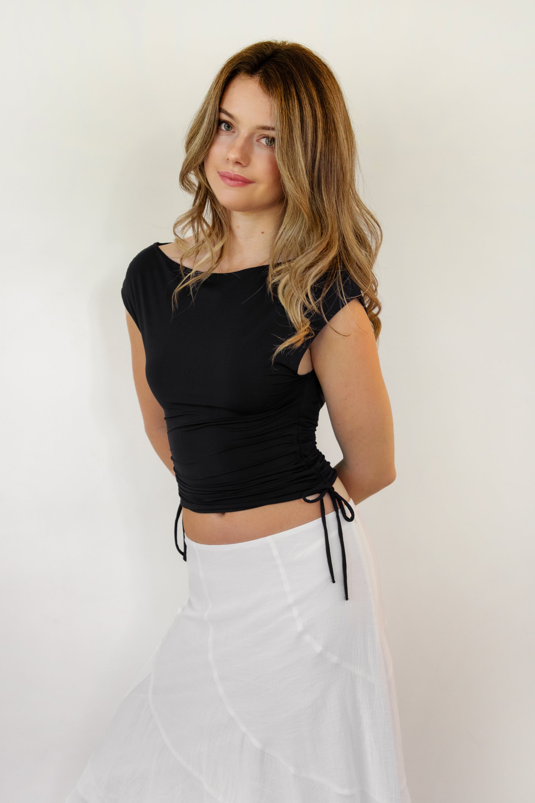 Woman wearing SELENE boat neck cowl back side tie crop top featuring an elegant boat neckline, draped cowl back, adjustable back strap, side tie ruching details, cap short sleeves, and lightweight stretch jersey fabric for a feminine cropped silhouette.