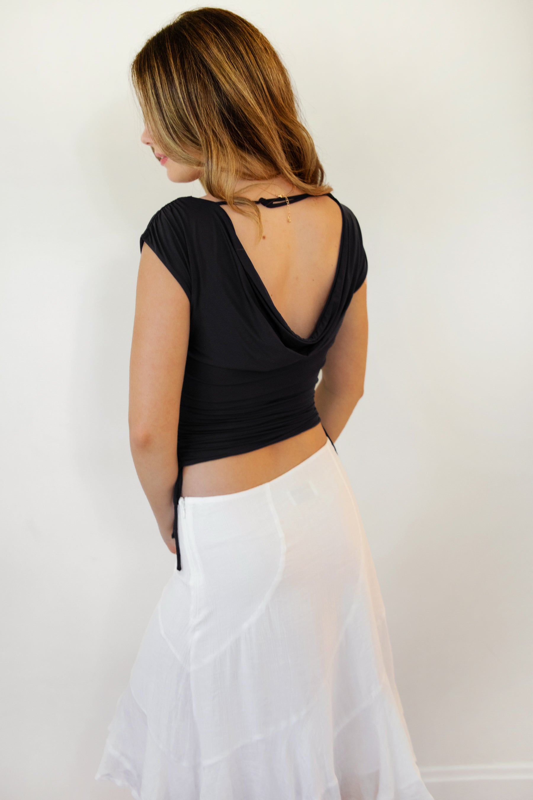 Woman wearing SELENE boat neck cowl back side tie crop top featuring an elegant boat neckline, draped cowl back, adjustable back strap, side tie ruching details, cap short sleeves, and lightweight stretch jersey fabric for a feminine cropped silhouette.