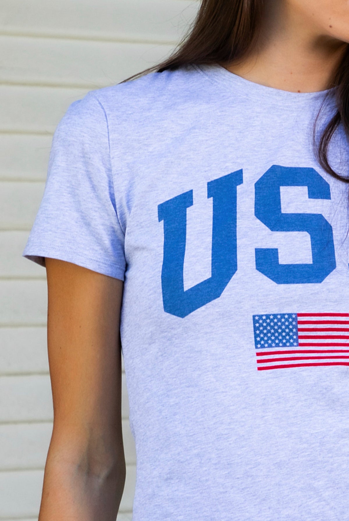 Woman wearing LIBERTY classic USA graphic crewneck baby tee featuring soft single jersey cotton stretch fabric, fitted short sleeve baby tee silhouette, clean cropped length, classic crew neckline, bold USA graphic across the front, American flag graphic detail below, and straight fitted hemline.