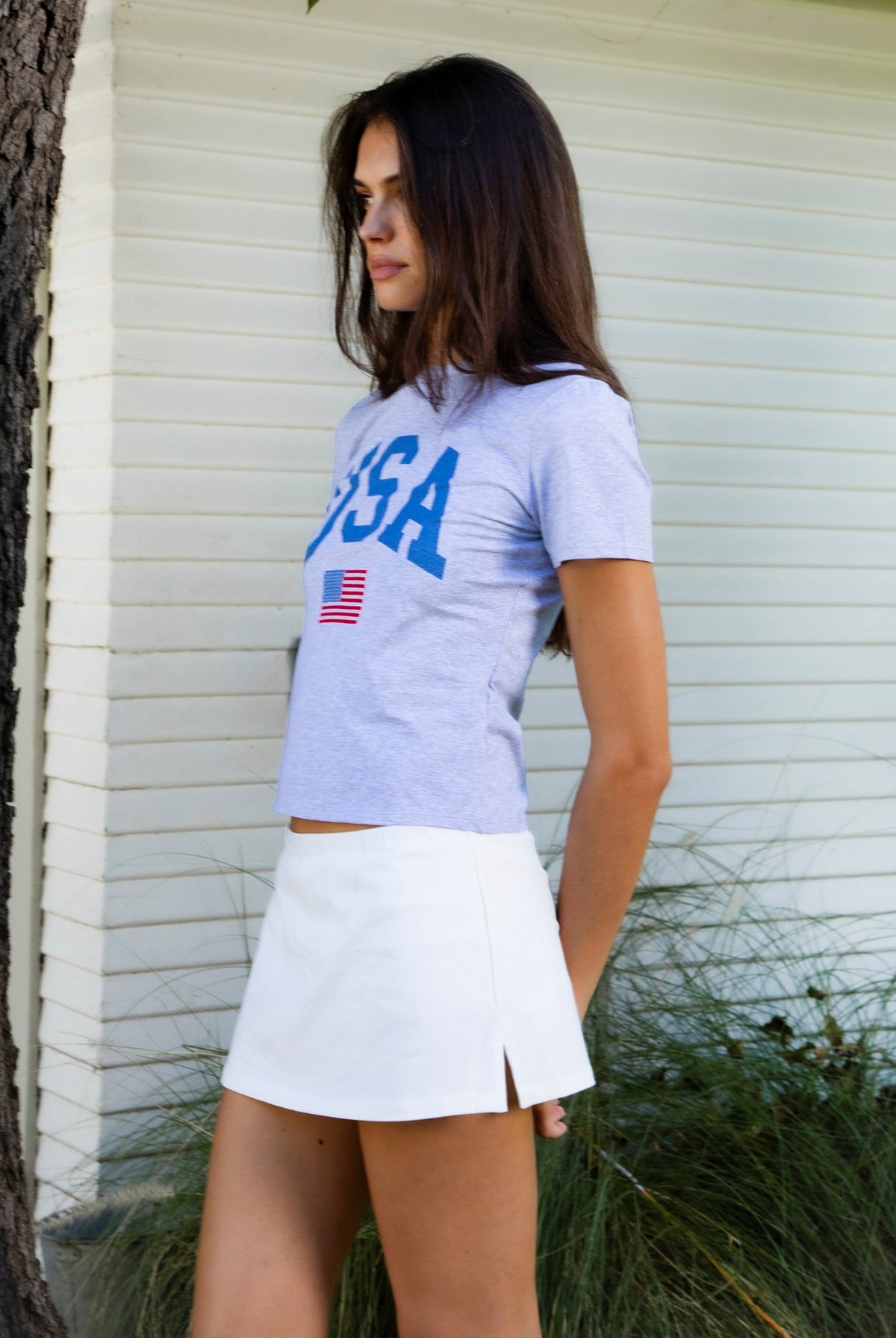 Woman wearing LIBERTY classic USA graphic crewneck baby tee featuring soft single jersey cotton stretch fabric, fitted short sleeve baby tee silhouette, clean cropped length, classic crew neckline, bold USA graphic across the front, American flag graphic detail below, and straight fitted hemline.